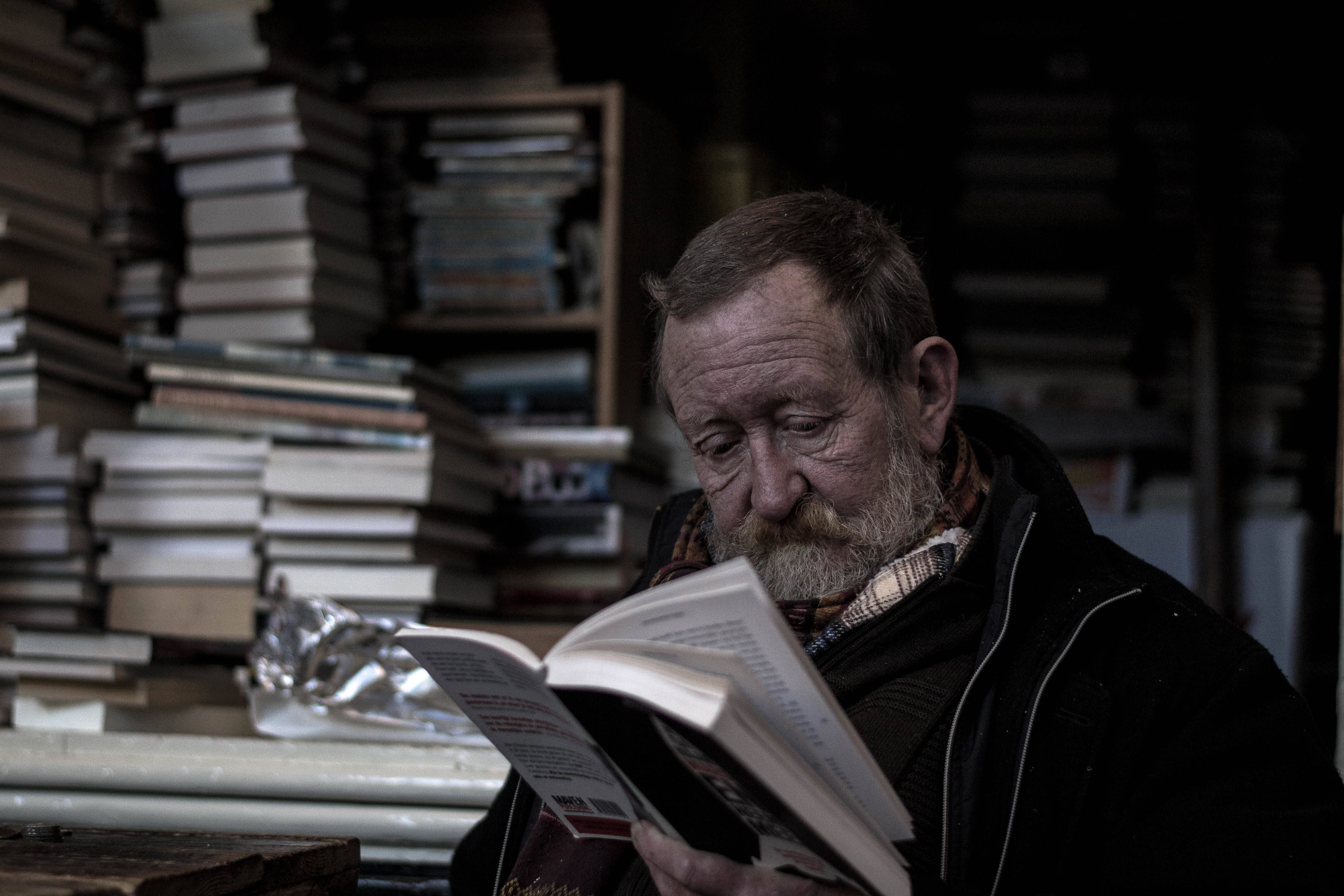 看書 桌布 壁紙 — 一位留鬍鬚的長者在一間燈光昏暗的房間裡，被高聳的書堆包圍著閱讀一本書。