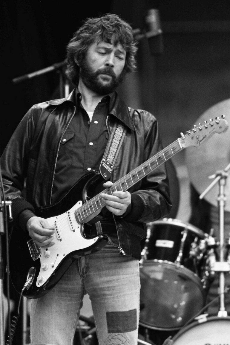guitar player 桌布 壁紙 — 一位留鬍鬚的音樂家穿著皮夾克，在舞台上熱情地彈奏白色電吉他，身後有鼓。