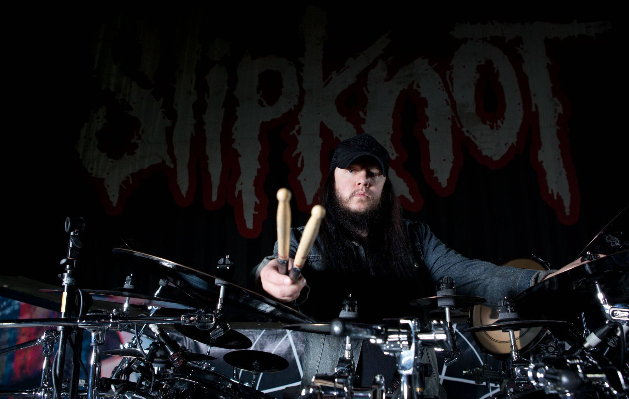 Joey Jordison 桌布 壁紙 — 一位留鬍鬚的鼓手戴著黑色帽子，在戲劇性的舞台燈光下，於金屬鼓組前手持鼓棒，身後是血紅色的Slipknot標誌。