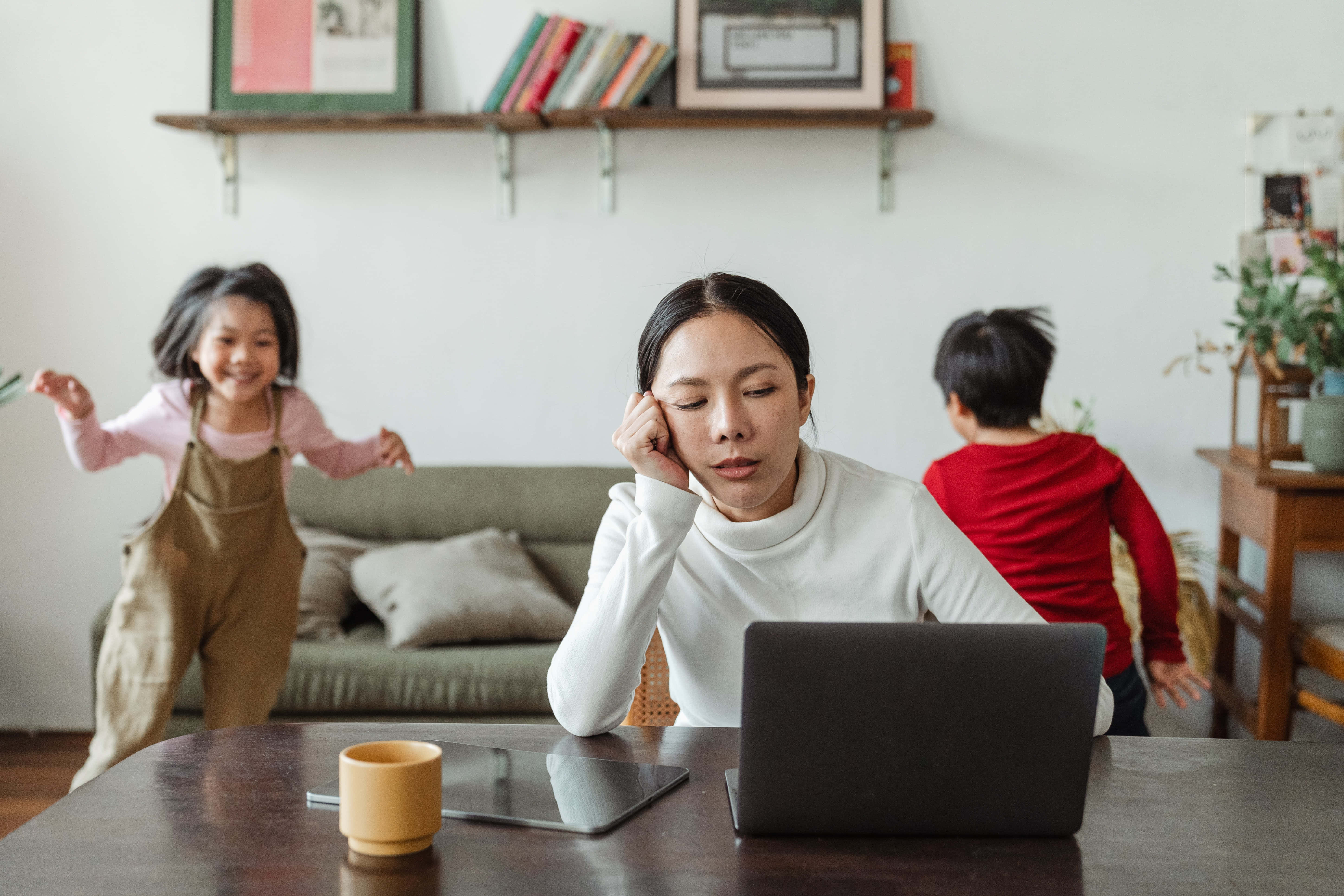 Mom 桌布 壁紙 — 一位疲倦的女士在工作時用手托著頭，兩個孩子在她身後明亮的家中精力充沛地玩耍。