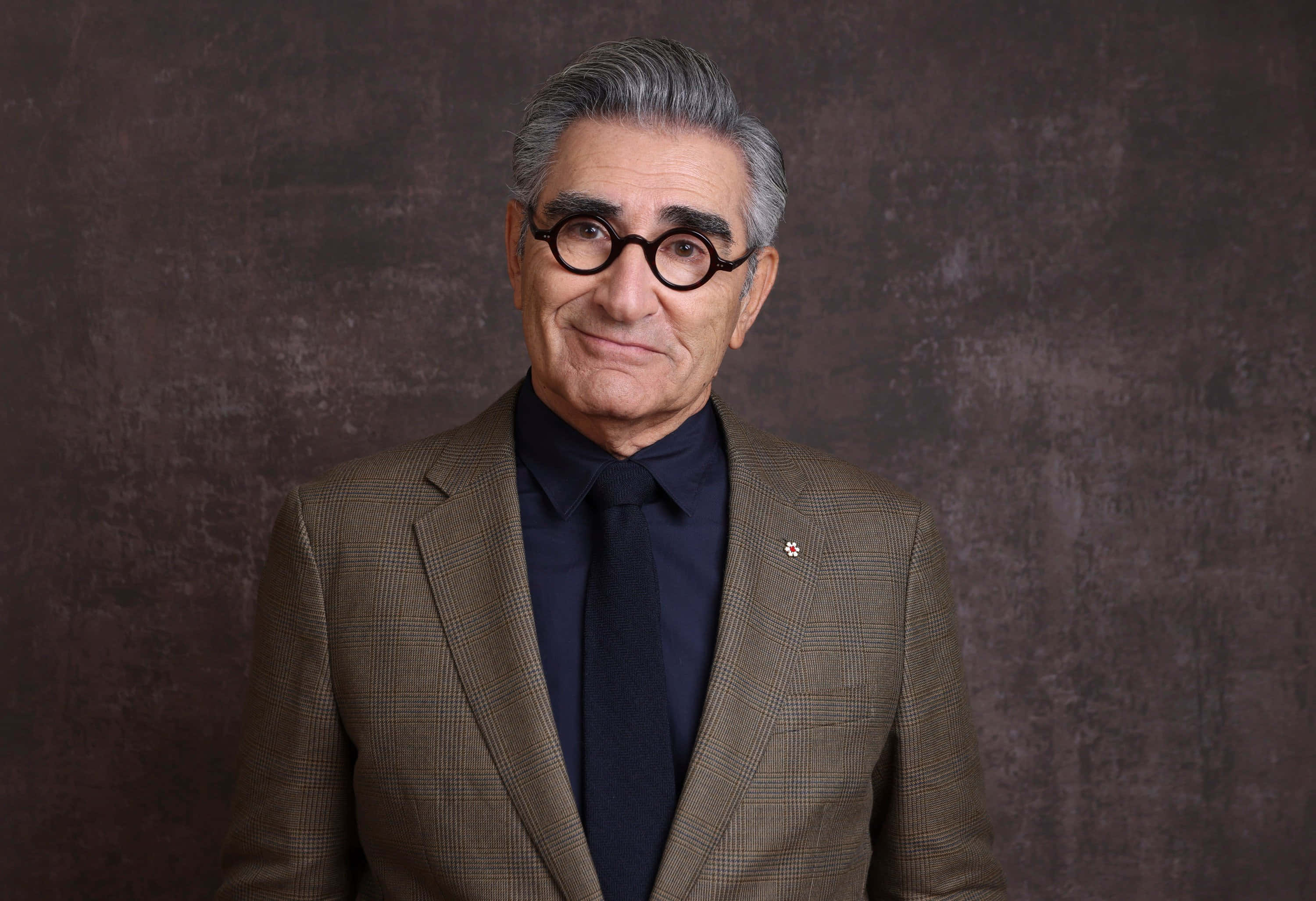 Eugene Levy 桌布 壁紙 — 一位白髮、戴圓框眼鏡的尊貴長者，身穿棕色格紋西裝，在帶有紋理的深棕色背景前溫暖地微笑。