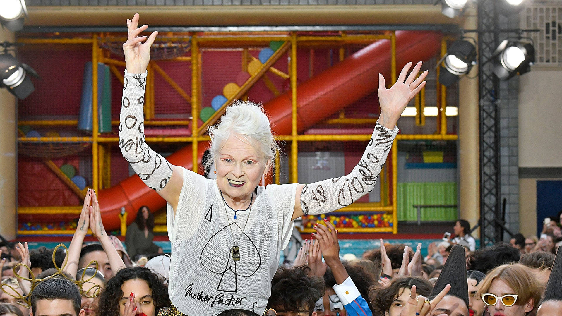 Vivienne Westwood 桌布 壁紙 — 一位白髮、手臂上畫有彩繪的老年女性，在一個充滿活力的室內活動中，高興地高舉雙手，高於歡呼的人群。