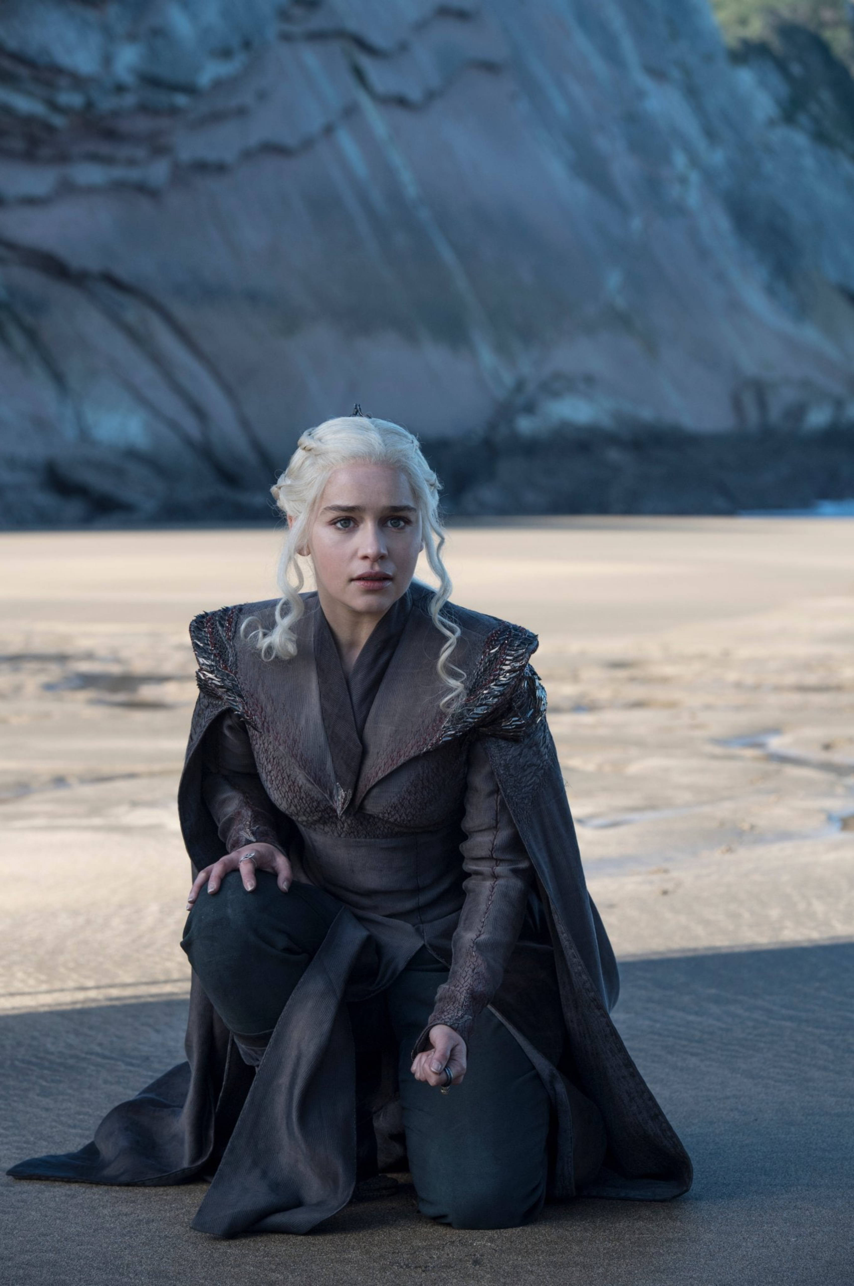 daenerys 桌布 壁紙 — 一位白髮女子在柔和日光下跪在沙灘上，面前是高聳的懸崖。