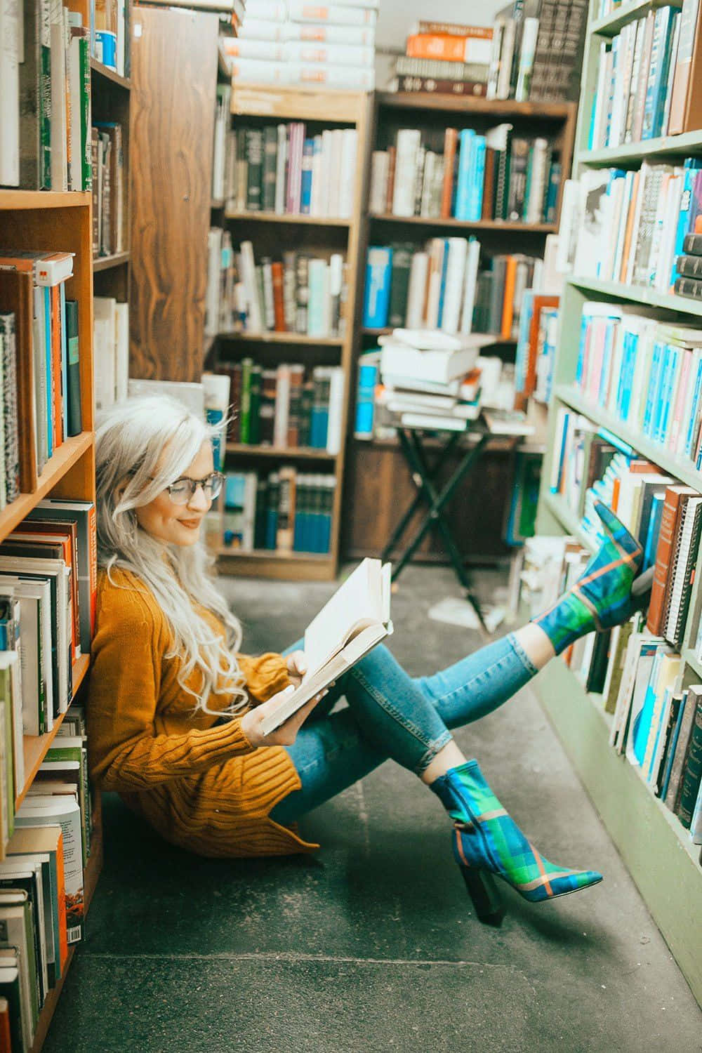 Library 桌布 壁紙 — 一位白髮女性盤腿坐在舒適圖書館的書架之間，穿著色彩繽紛的格紋靴子閱讀一本書。