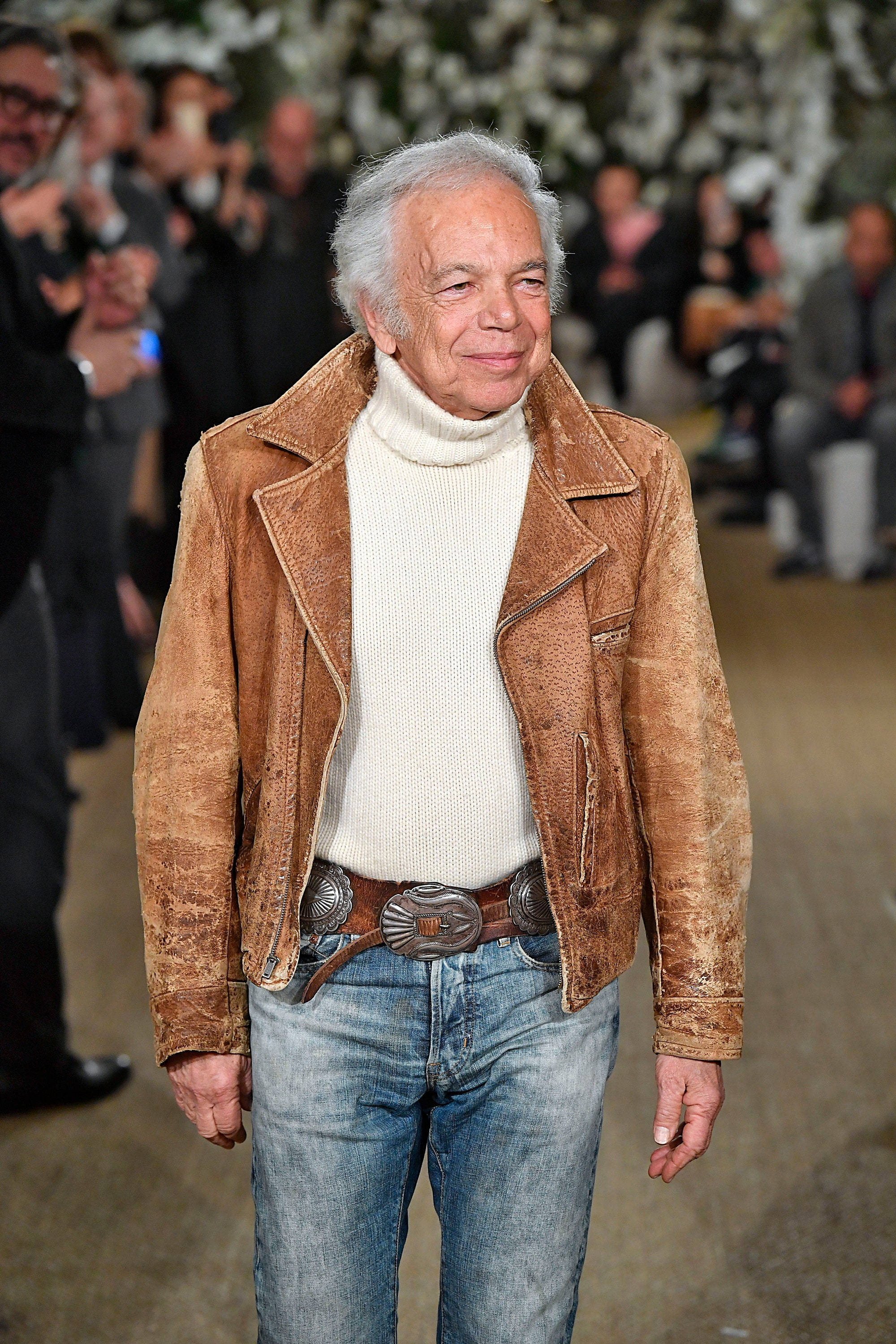 ralph lauren 桌布 壁紙 — 一位白髮年長男士穿著棕色麂皮外套內搭白色高領毛衣和牛仔褲微笑。