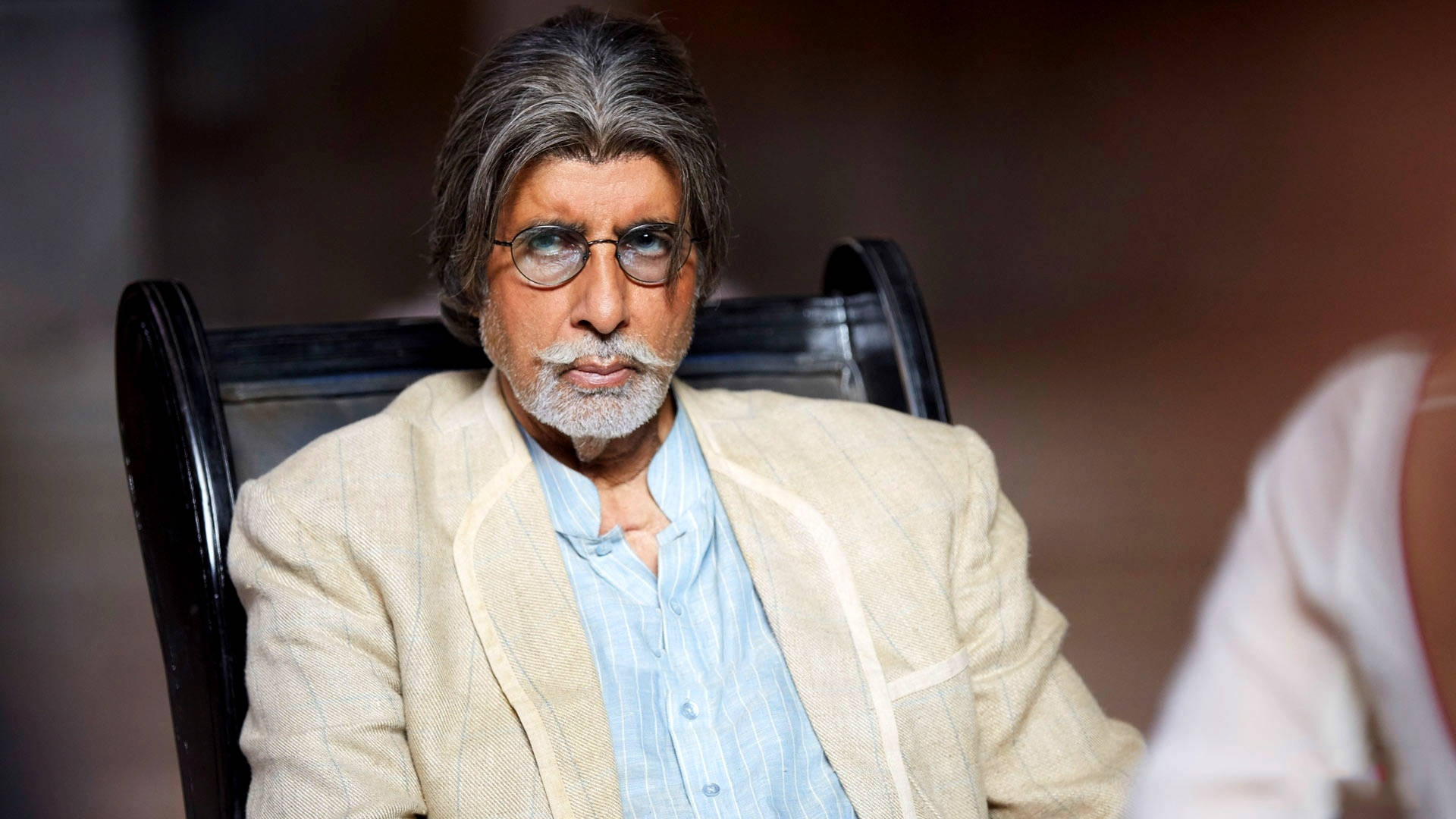Amitabh Bachchan 桌布 壁紙 — 一位白髮戴眼鏡的尊貴長者坐在深色木椅上，身穿米色西裝外套，內搭淺藍色襯衫。