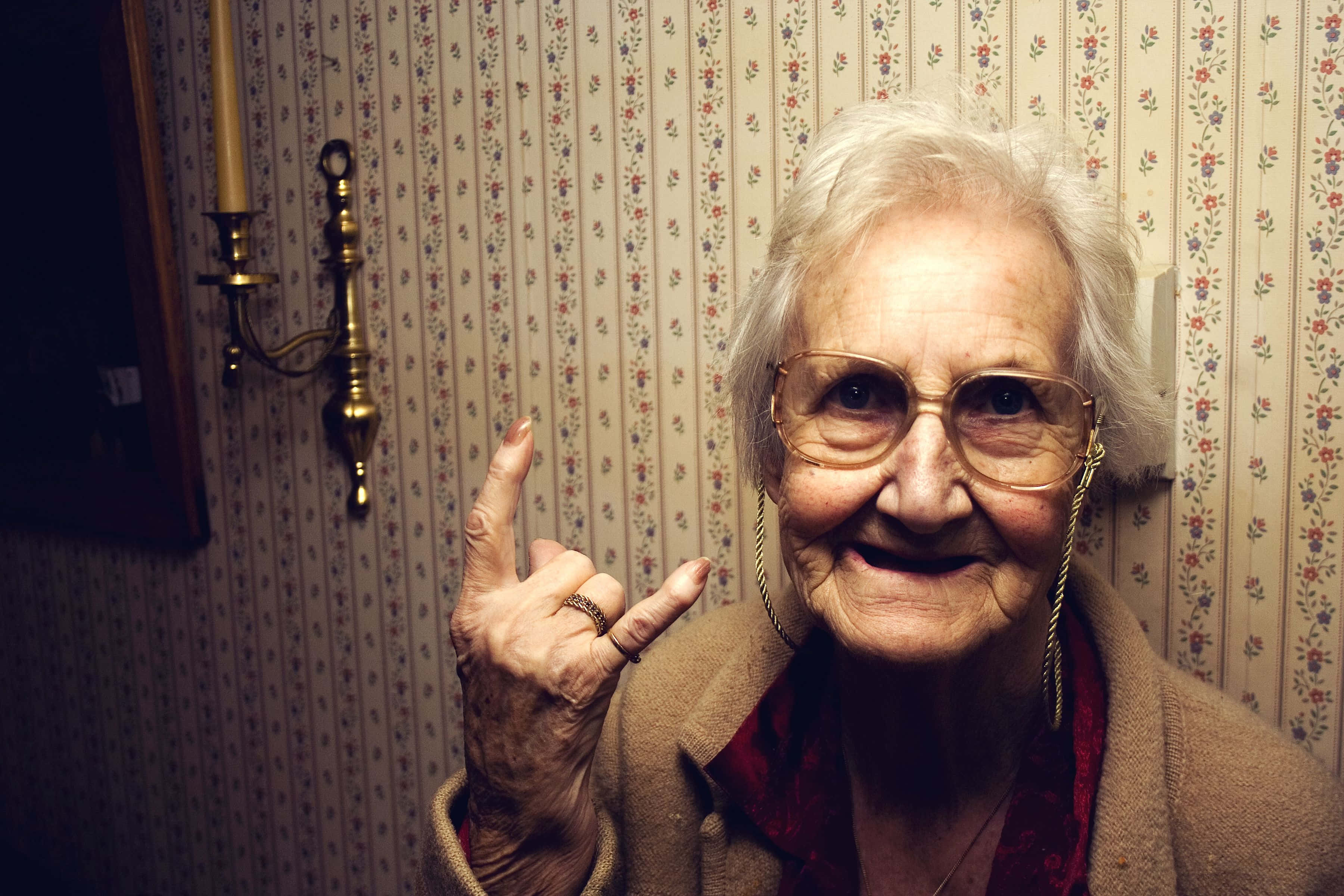 Old Lady 桌布 壁紙 — 一位白髮戴眼鏡的老婦人，在花卉壁紙前微笑著做出搖滾手勢。