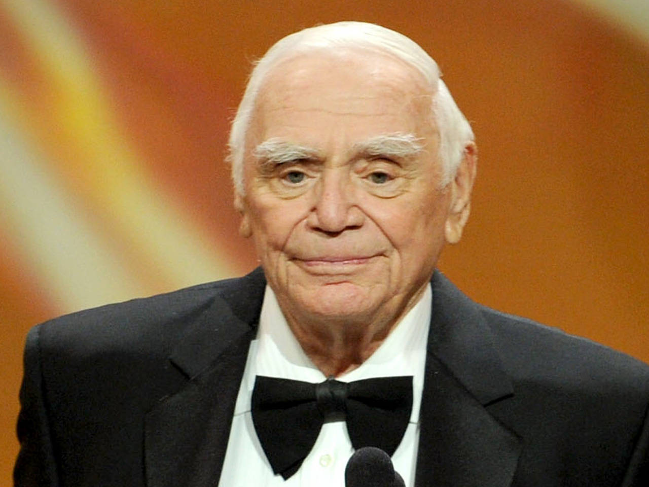 actor 桌布 壁紙 — 一位白髮戴領結的長者，在溫暖燈光的舞台上，站在麥克風前露出溫暖的笑容。