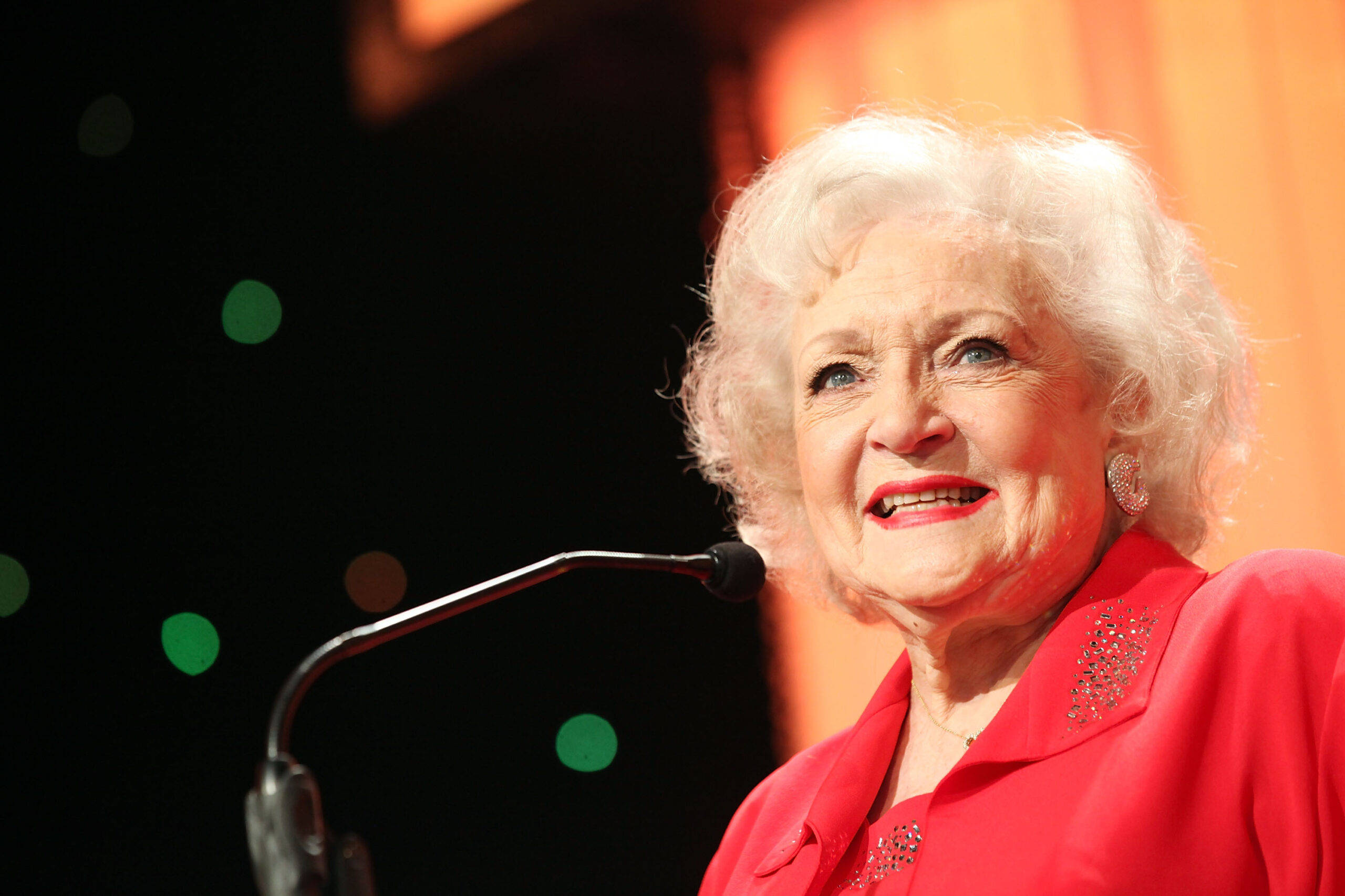 Betty White 桌布 壁紙 — 一位白髮捲髮、塗著紅色口紅的老年女性，在橙色和綠色燈光的舞台上對著麥克風溫暖地微笑說話。
