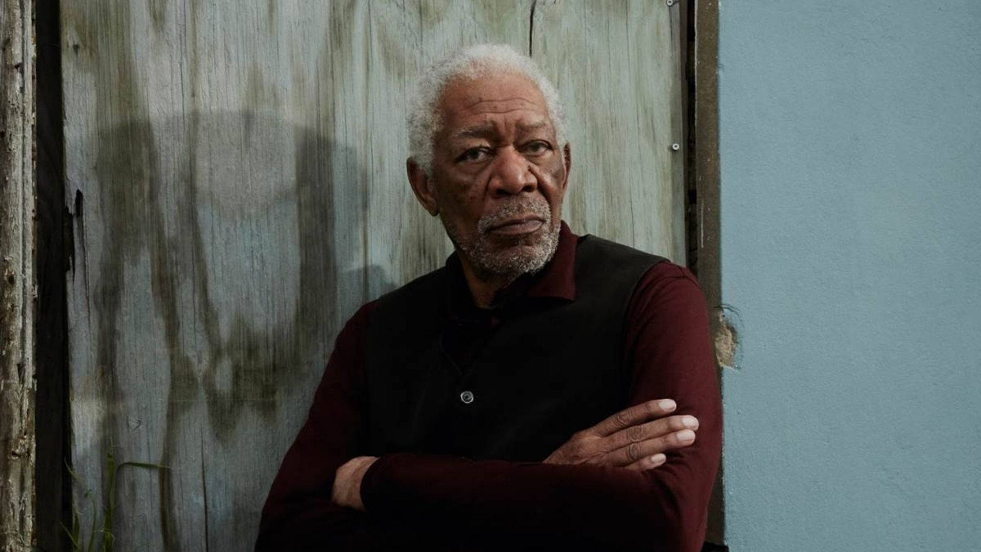Morgan Freeman 桌布 壁紙 — 一位白髮白鬍子的長者雙臂交叉，站在風化的木牆和藍色漆面背景前。