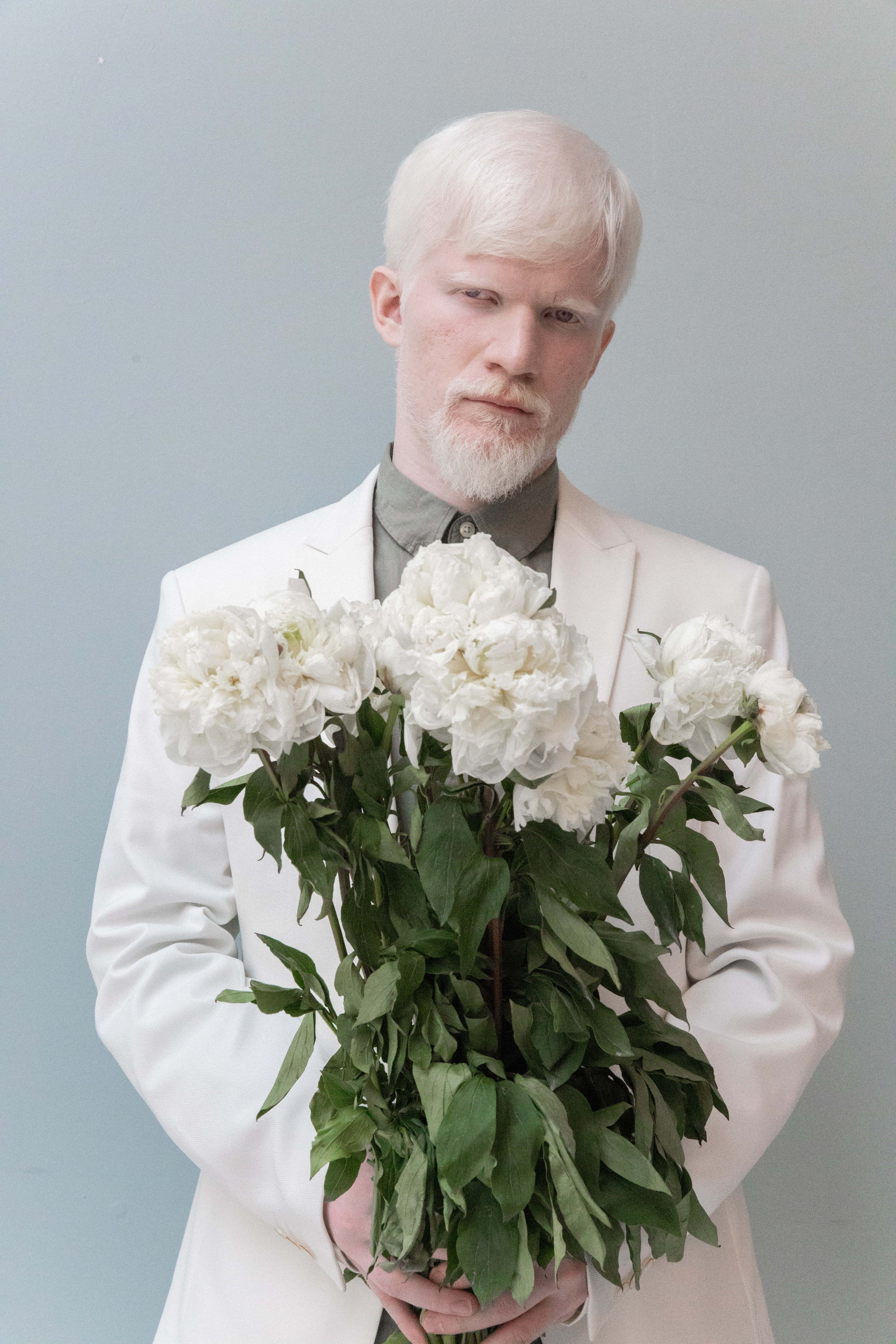 Man In Suit 桌布 壁紙 — 一位白髮白鬍鬚的人穿著白色西裝，在柔和的灰色背景前拿著一大束白色牡丹花。