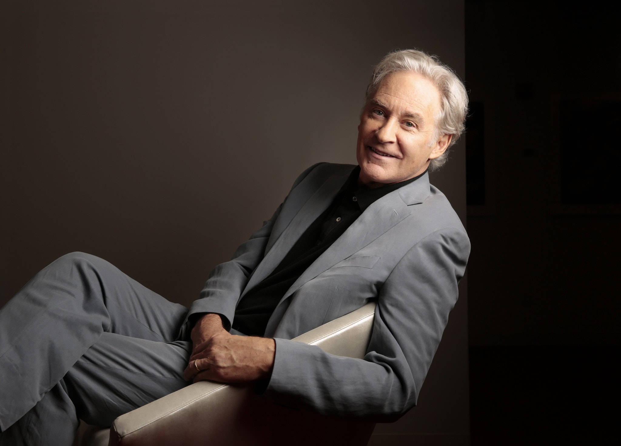 攝影 桌布 壁紙 — 一位白髮的尊貴長者，在深色攝影棚背景前，現代椅子上悠閒地微笑。