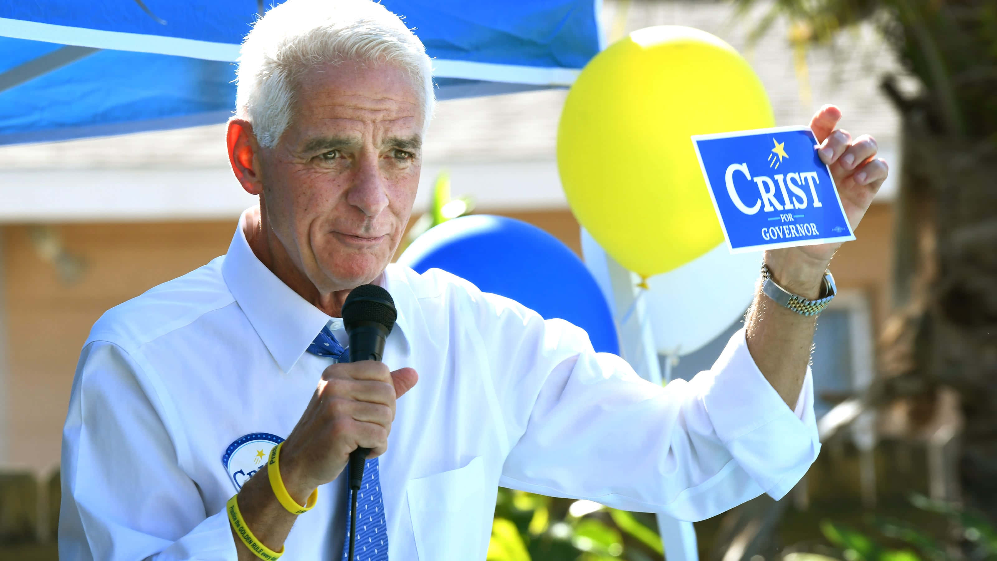 桌布 壁紙 — 一位白髮老人在戶外競選活動中，手持藍色「Crist for Governor」標語牌，對著麥克風講話。