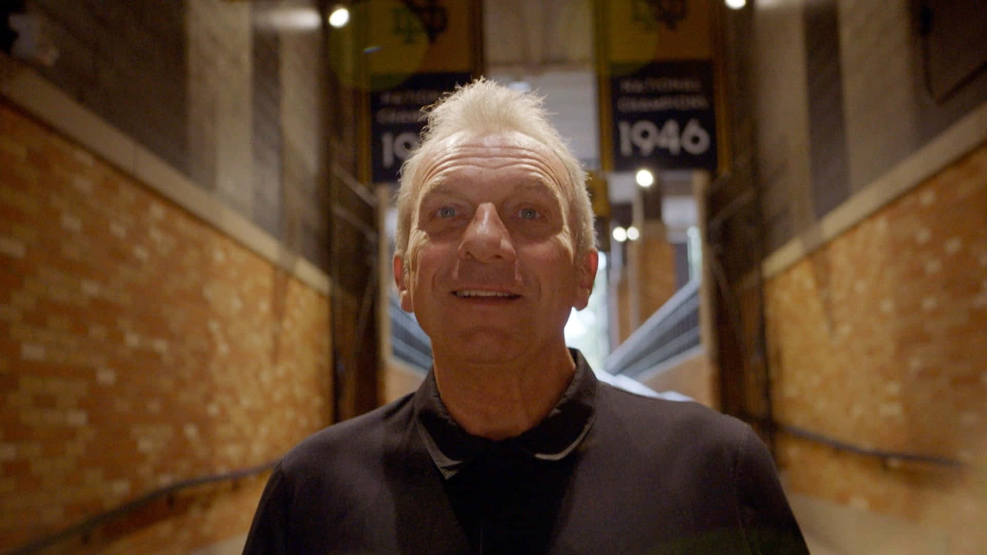 Joe Montana 桌布 壁紙 — 一位白髮老人在燈光昏暗的磚砌走廊中微笑，走廊裝飾著冠軍錦旗。