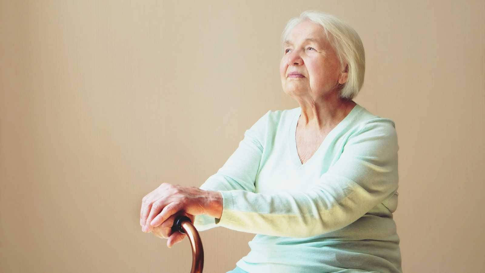 Sitting Alone 桌布 壁紙 — 一位白髮老婦人平靜地坐著，手持木製拐杖，背景是柔和的米色牆壁。