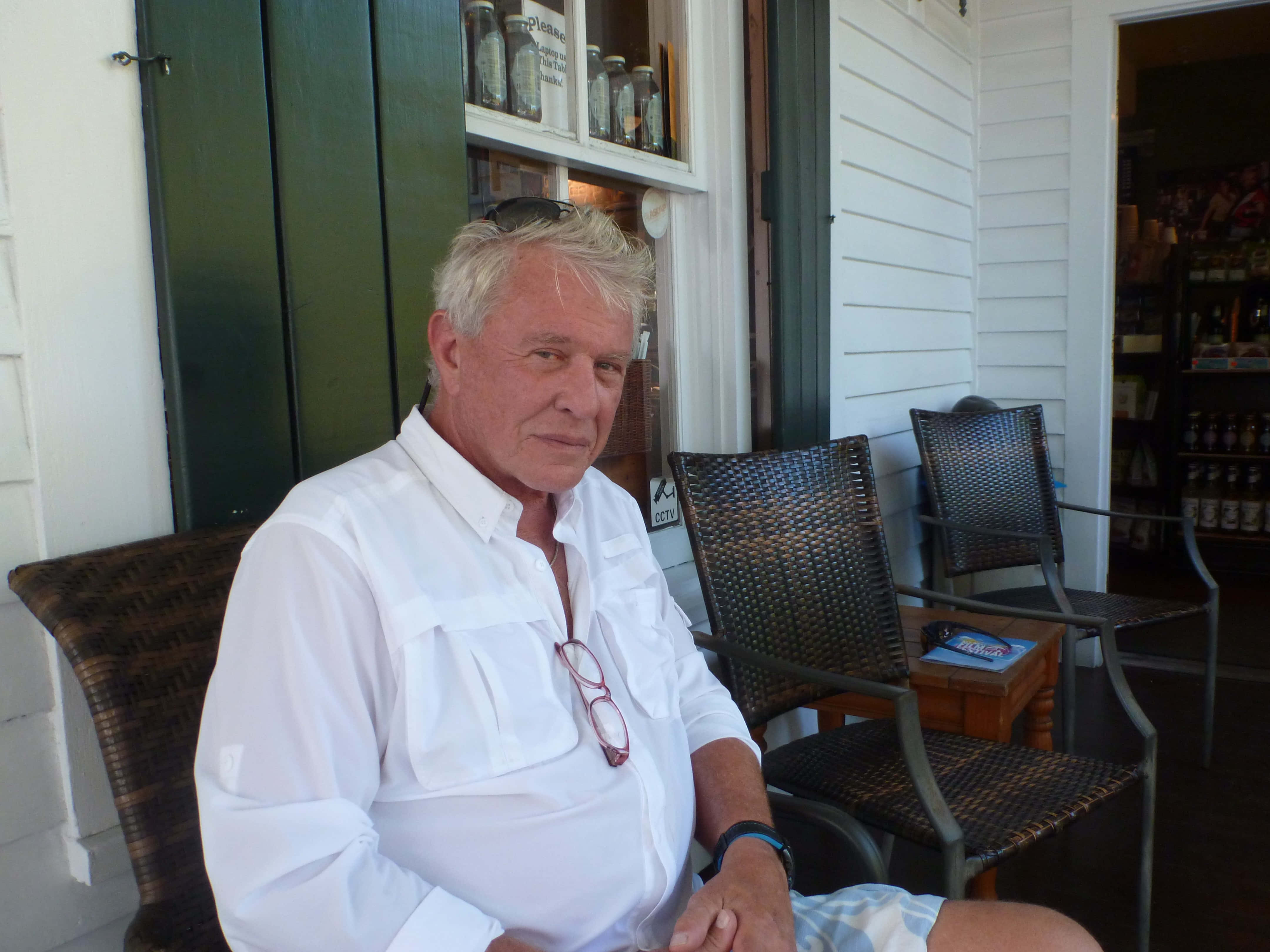 Tom Berenger 桌布 壁紙 — 一位白髮老翁平靜地坐在門廊的藤椅上，身穿白襯衫，眼鏡掛在衣領上。