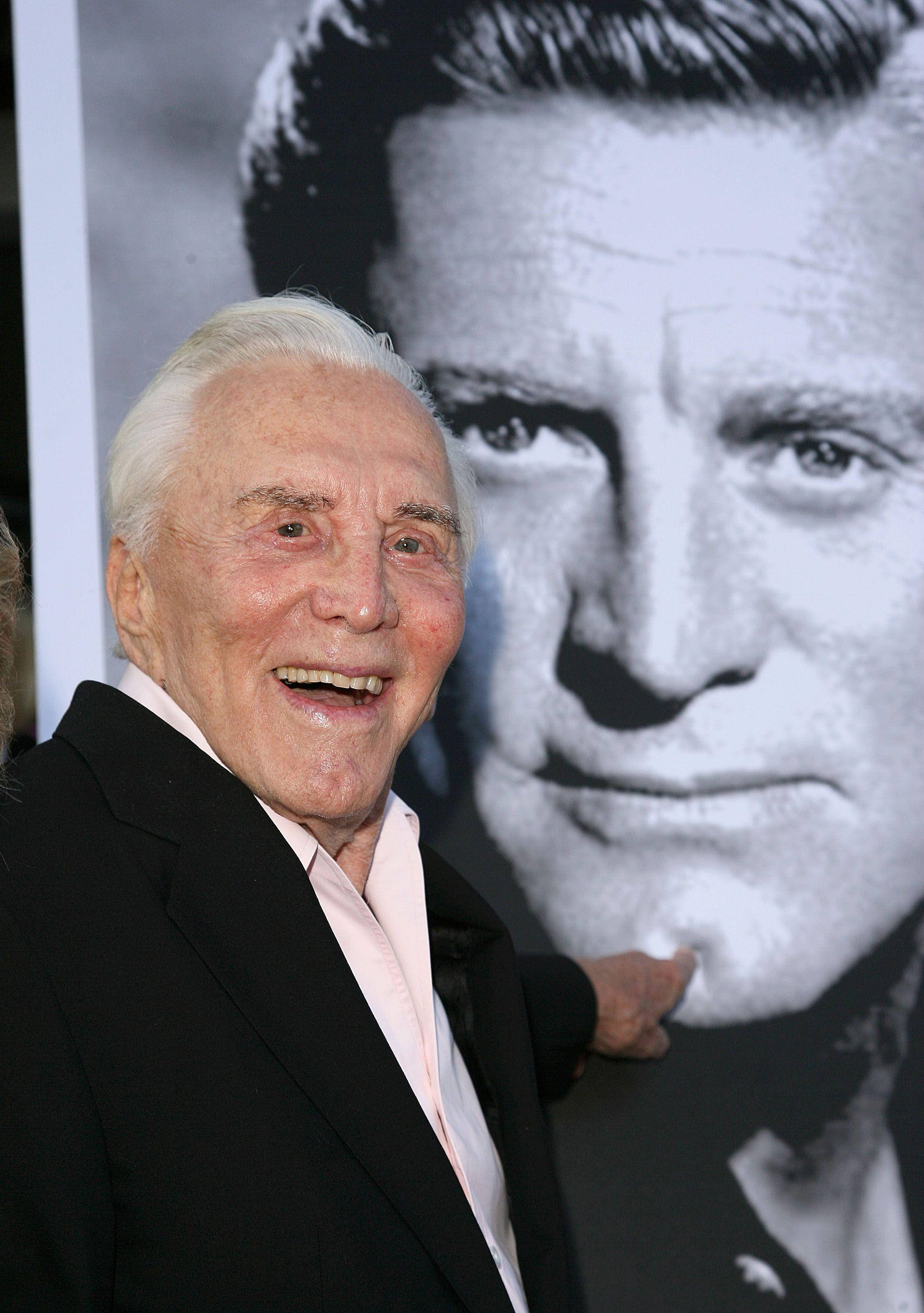 actor 桌布 壁紙 — 一位白髮老翁身穿黑色西裝，在一個年輕男子的黑白大畫像旁露出燦爛笑容。