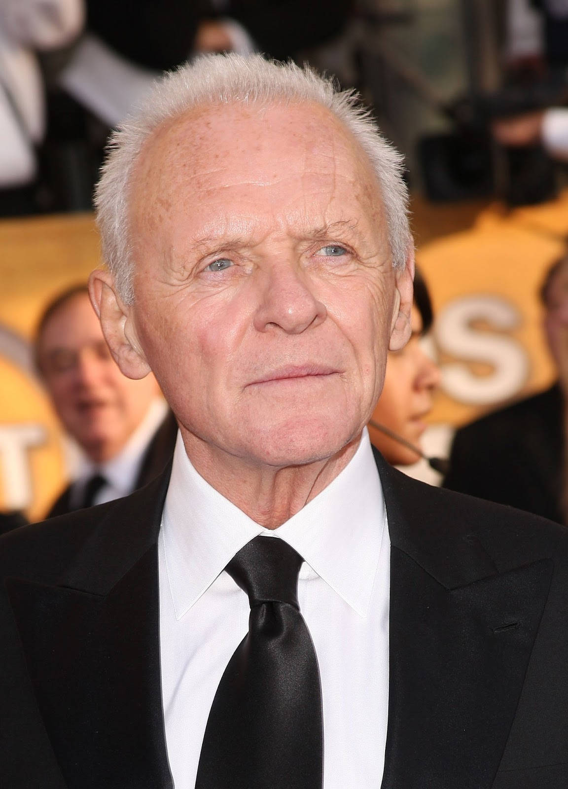 actor 桌布 壁紙 — 一位白髮藍眼的年長男士在模糊背景的正式活動中穿著黑色西裝打領帶。