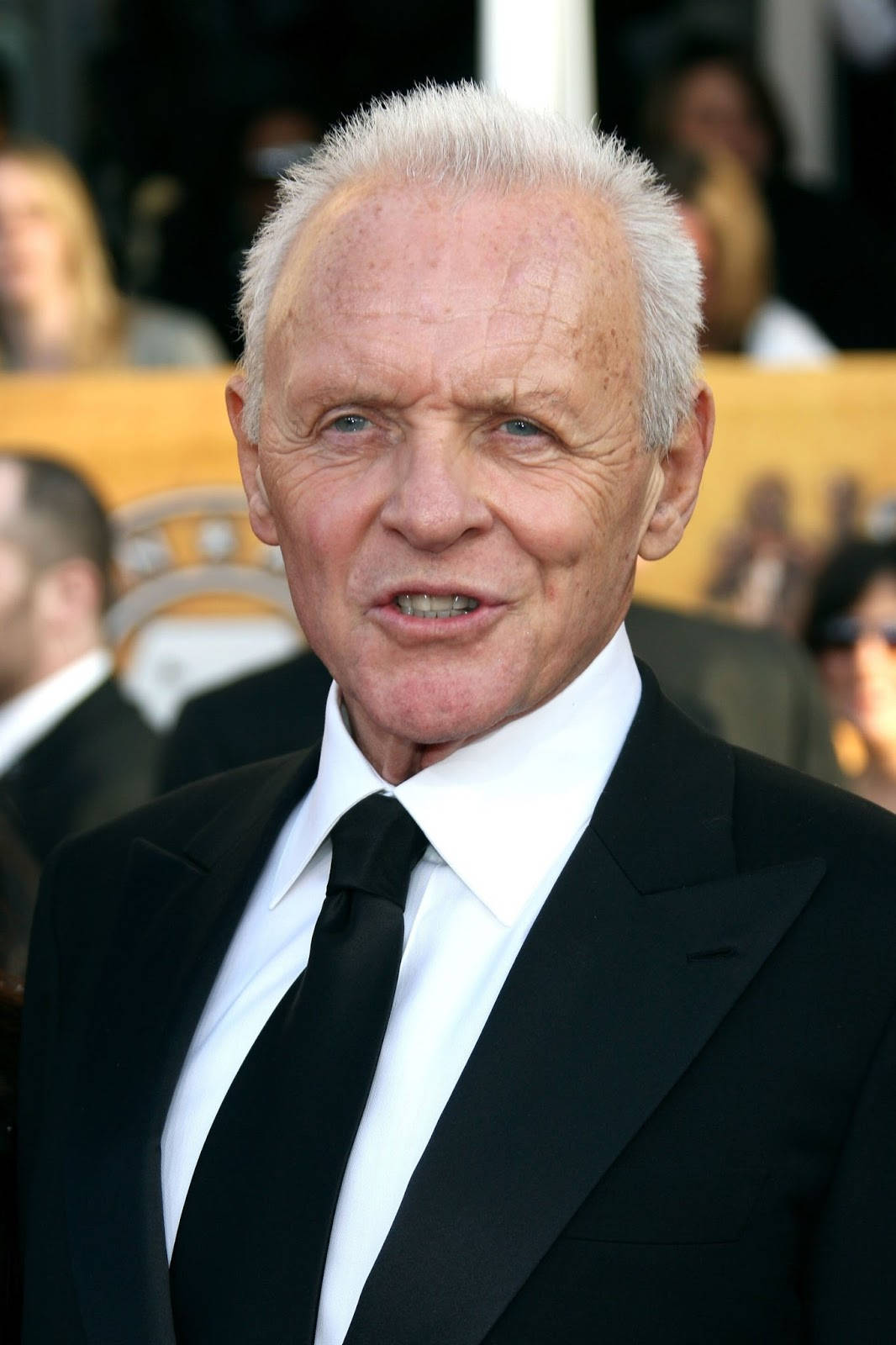 actor 桌布 壁紙 — 一位白髮藍眼的年長男士在正式活動中穿著黑色西裝和領帶，背景人物模糊。