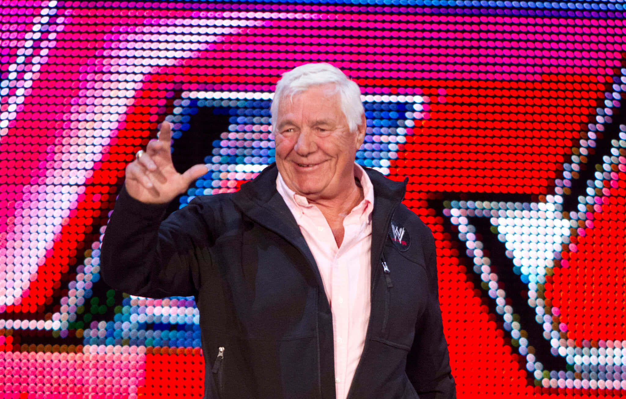 WWE 桌布 壁紙 — 一位白髮長者在充滿活力的像素化WWE標誌背景前開心地揮手。
