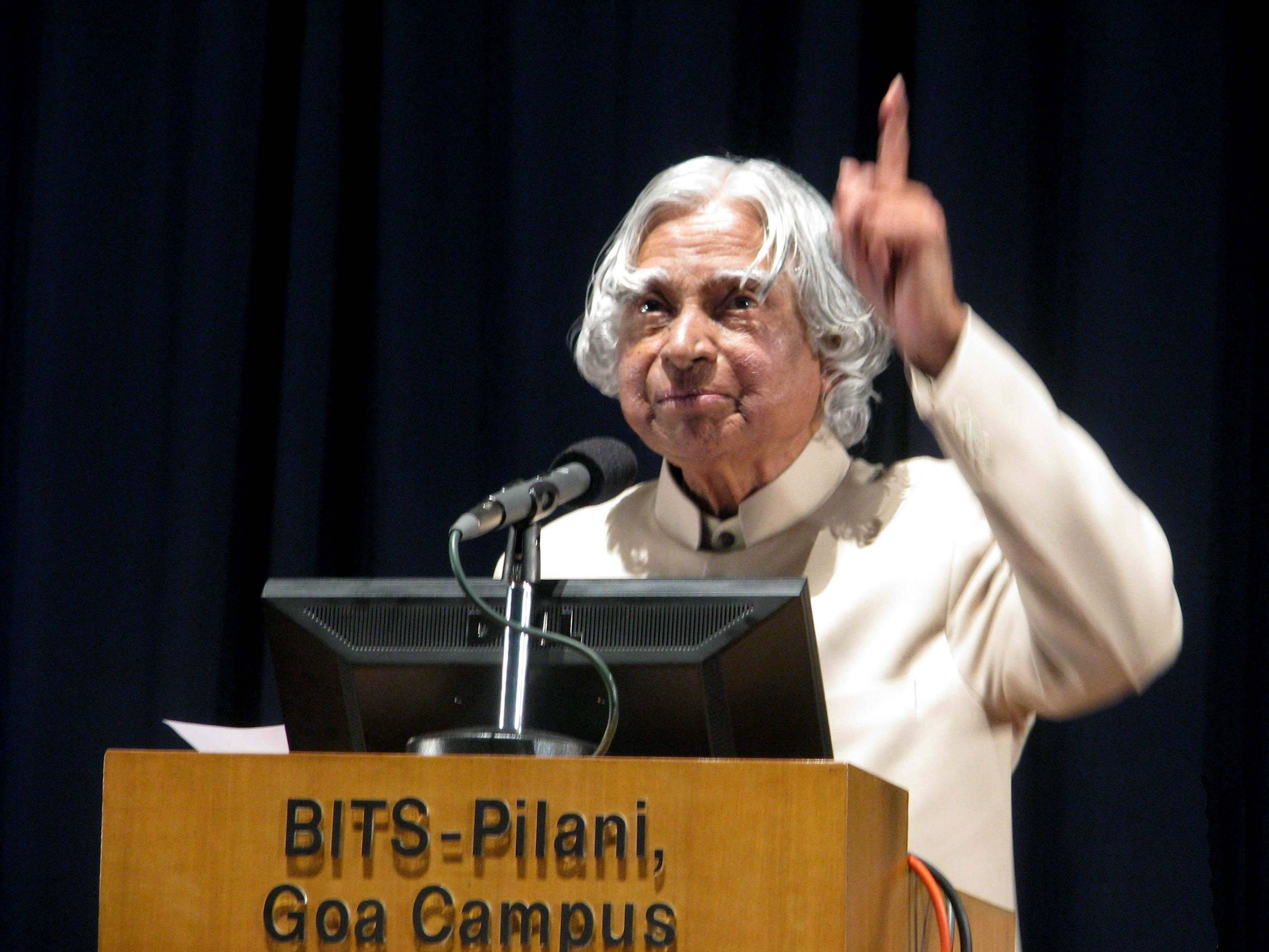 政治 桌布 壁紙 — 一位白髮長者在標有BITS-Pilani Goa Campus的講台上，右手向上揮動，充滿熱情地演講。