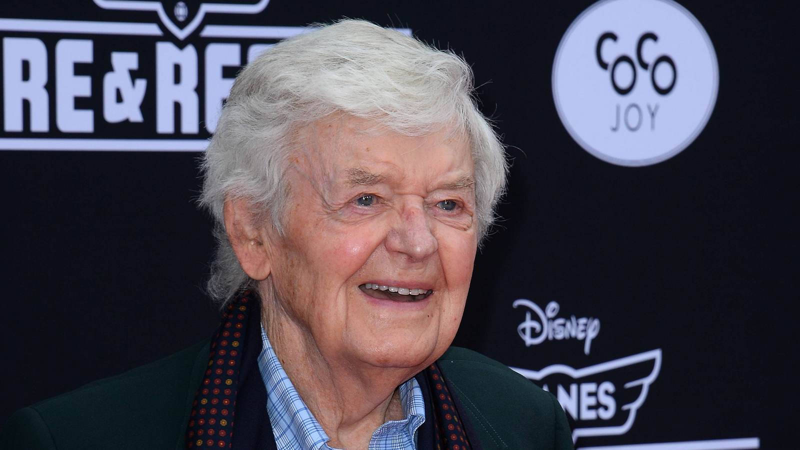 Celebrity 桌布 壁紙 — 一位白髮長者在迪士尼活動背景前，帶著Coco Joy和Re&Re標誌，露出溫暖笑容。