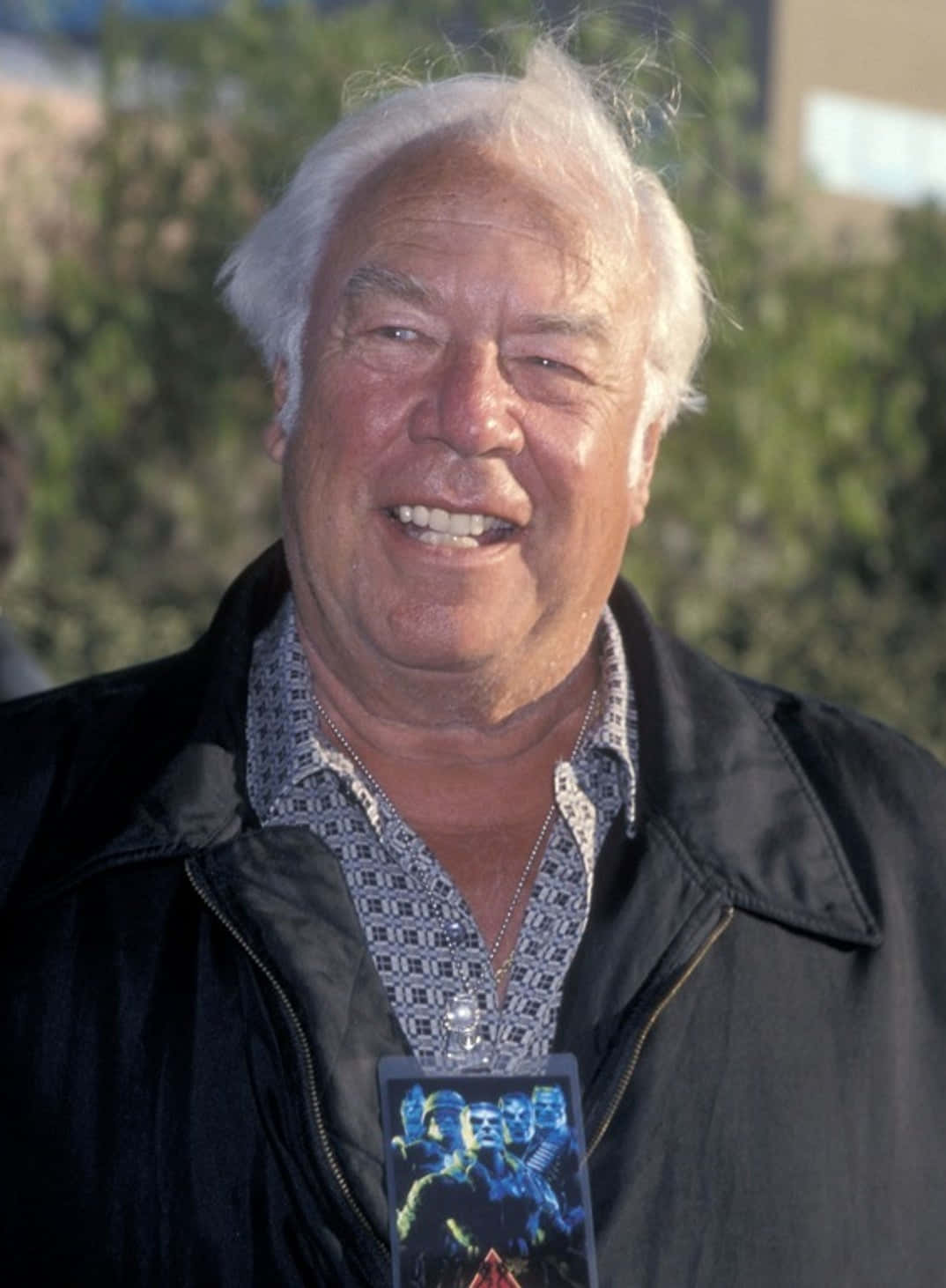 George Kennedy 桌布 壁紙 — 一位白髮長者穿著黑色外套，掛著電影海報掛繩，露出溫暖的笑容。