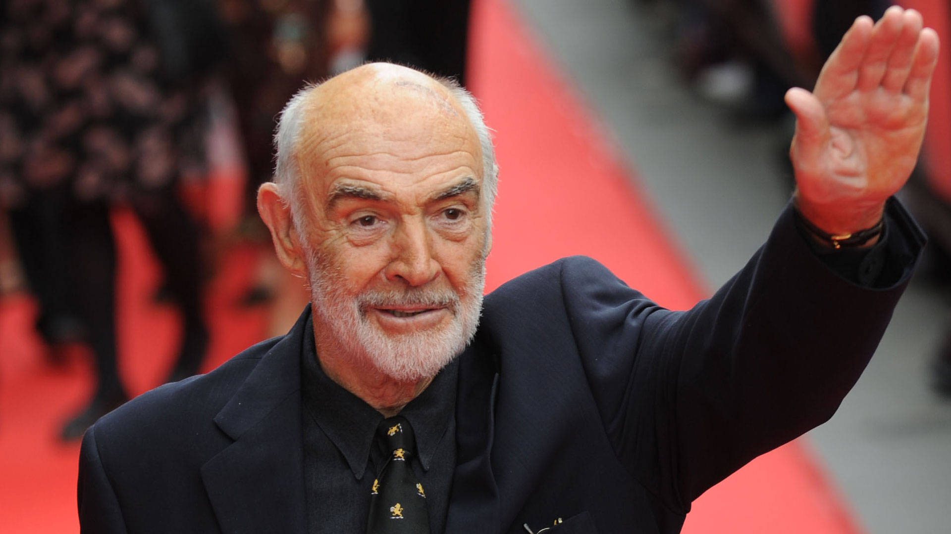 Sean Connery 桌布 壁紙 — 一位白鬚的尊貴長者身穿深色西裝，在紅地毯活動上熱情揮手。
