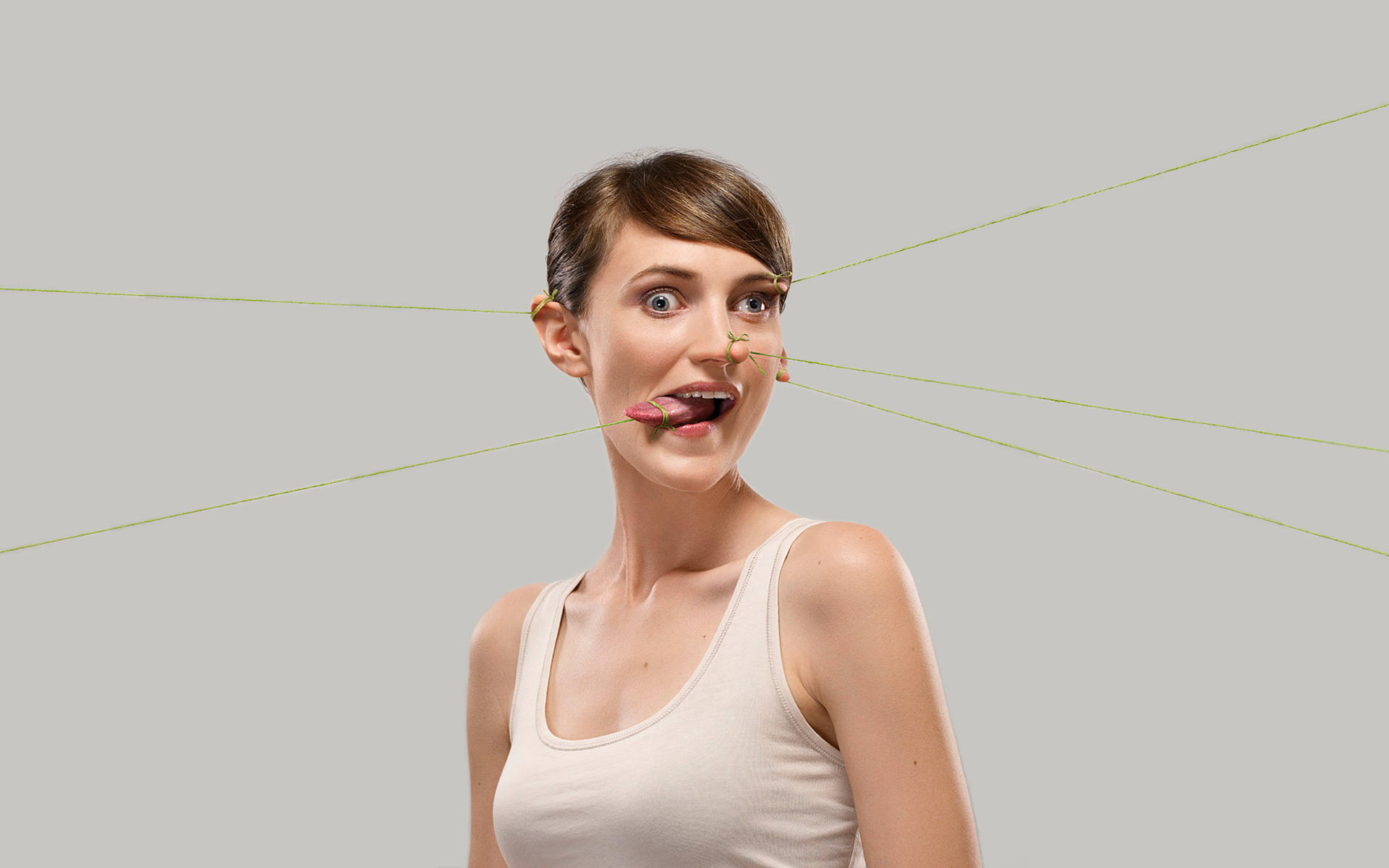 faces 桌布 壁紙 — 一位眼睛睜大、嘴巴張開的女性，臉上繫著細綠色的繩子，在純灰色背景上拉緊。