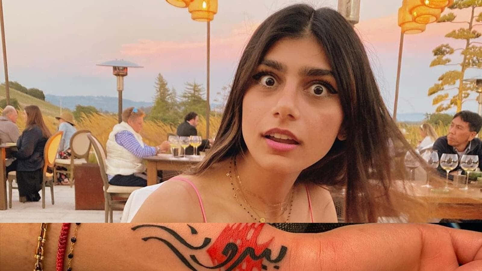Mia Khalifa 桌布 壁紙 — 一位眼睛睜大、表情驚訝的女性在日落戶外餐廳舉起手臂，展示紅色和黑色的刺青。
