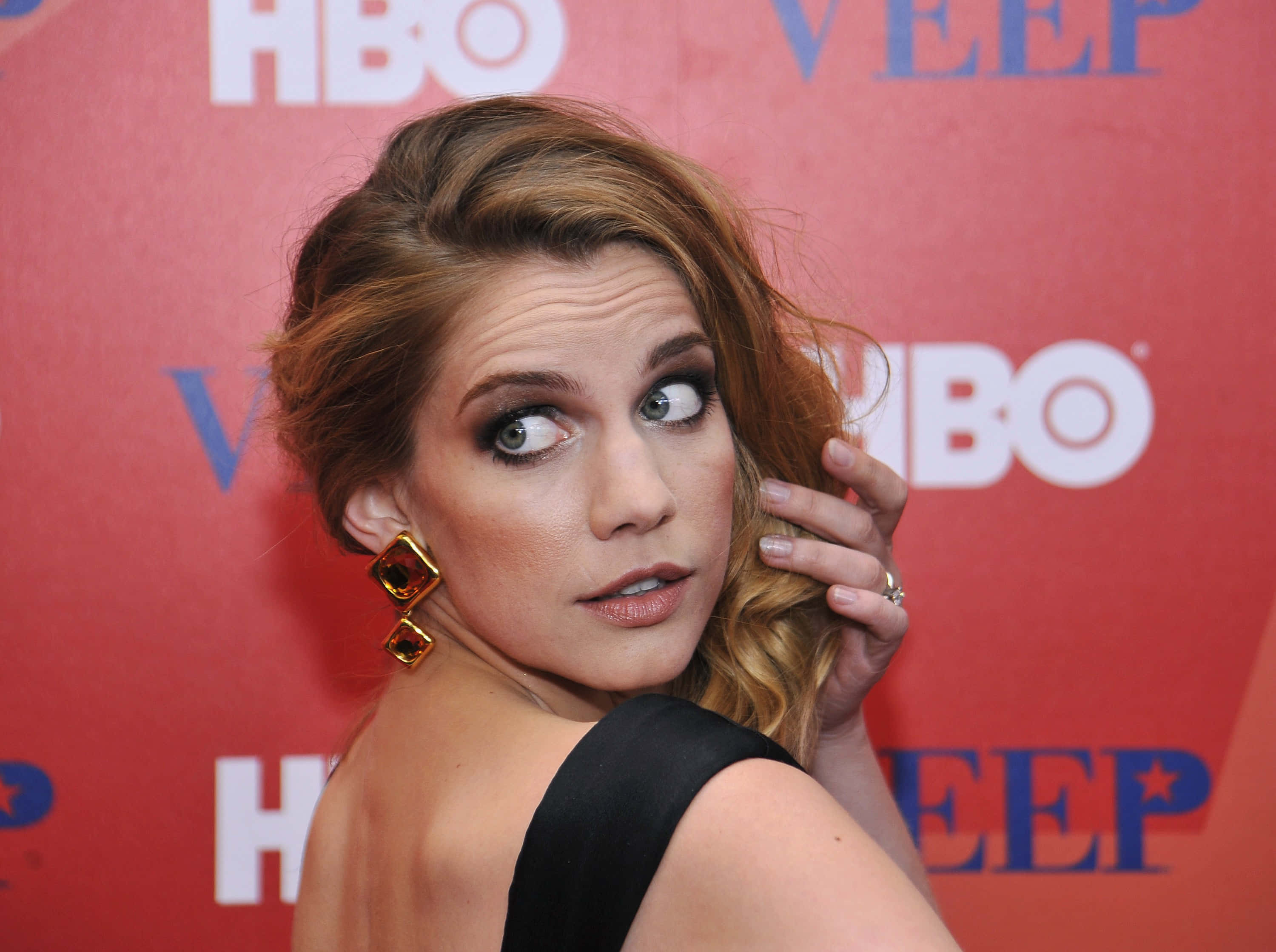 anna chlumsky 桌布 壁紙 — 一位眼神充滿表情、戴著大膽耳環的女性，在紅色HBO Veep背景前俏皮地擺姿勢。