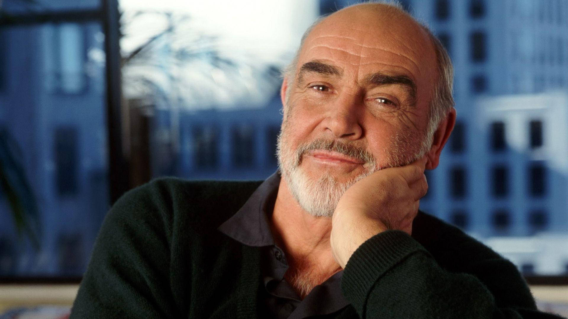 Sean Connery 桌布 壁紙 — 一位睿智的白鬍子長者，托著下巴，對著模糊的城市景觀窗戶，若有所思地凝視著相機。