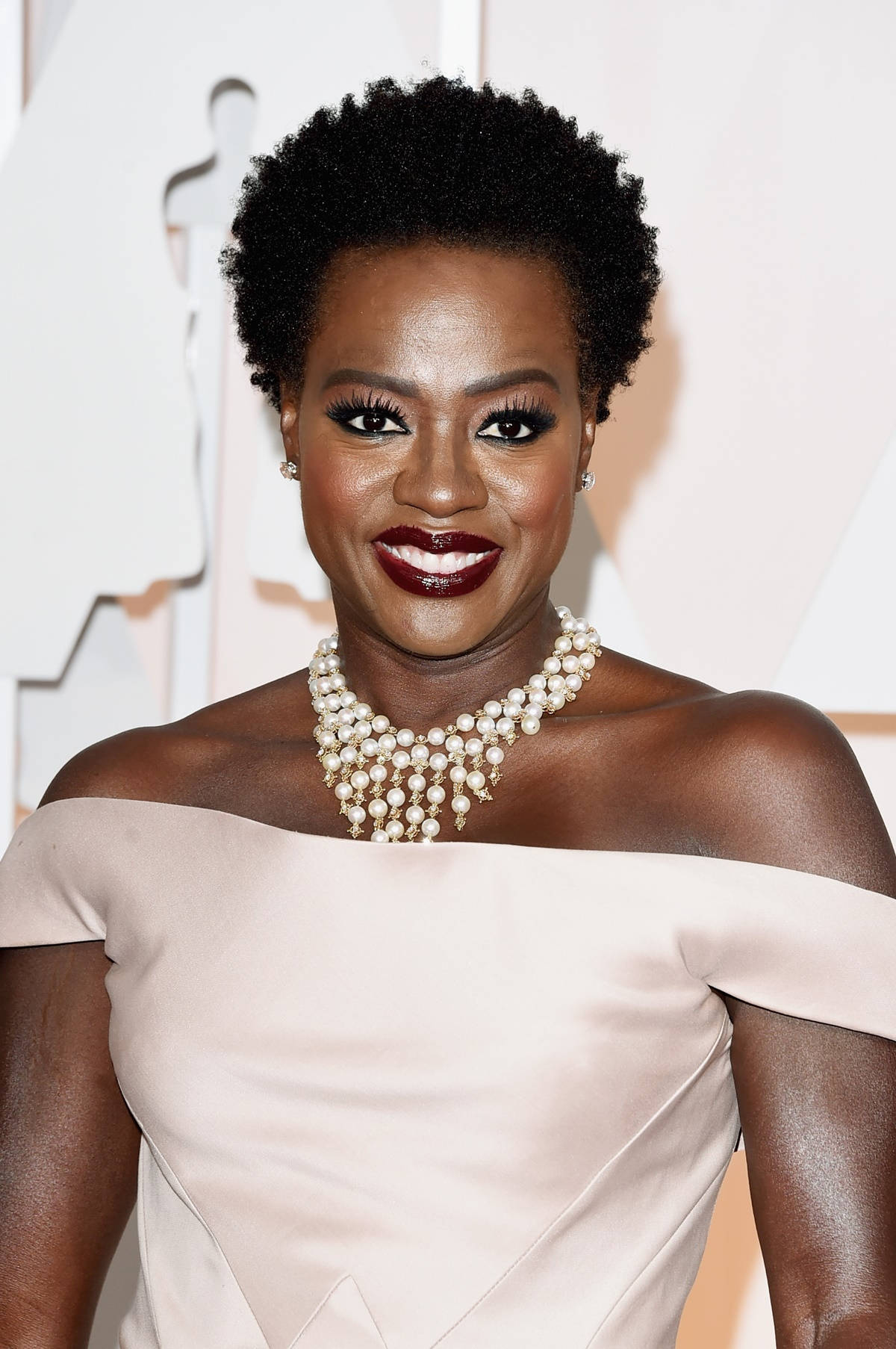 oscars 桌布 壁紙 — 一位短捲髮的燦爛女性，戴著珍珠項鍊，穿著露肩淡粉紅禮服，在華麗活動中微笑。