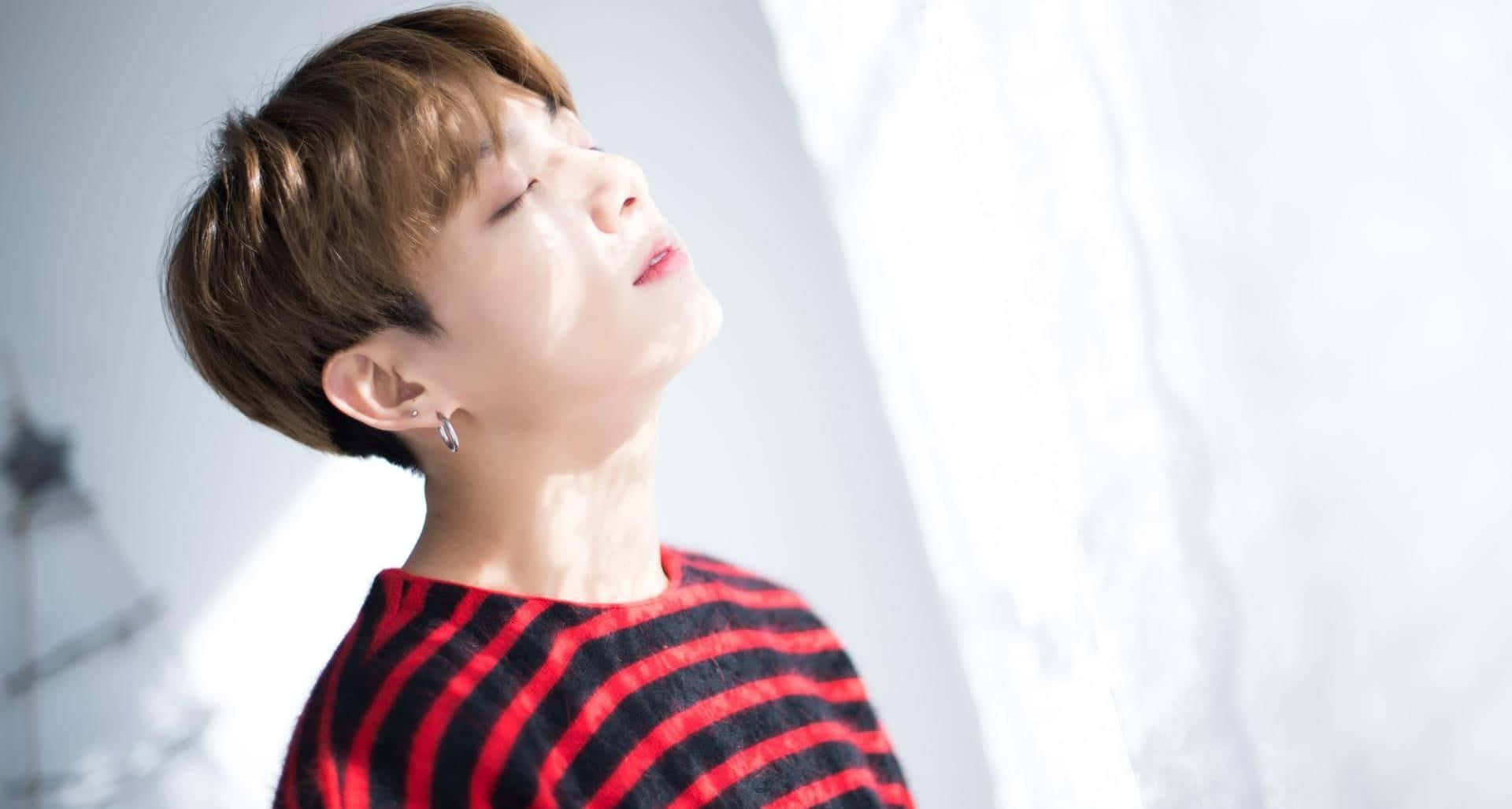 Jungkook 桌布 壁紙 — 一位短棕髮、身穿紅條紋毛衣的年輕人在窗邊柔和的陽光下向後仰頭。