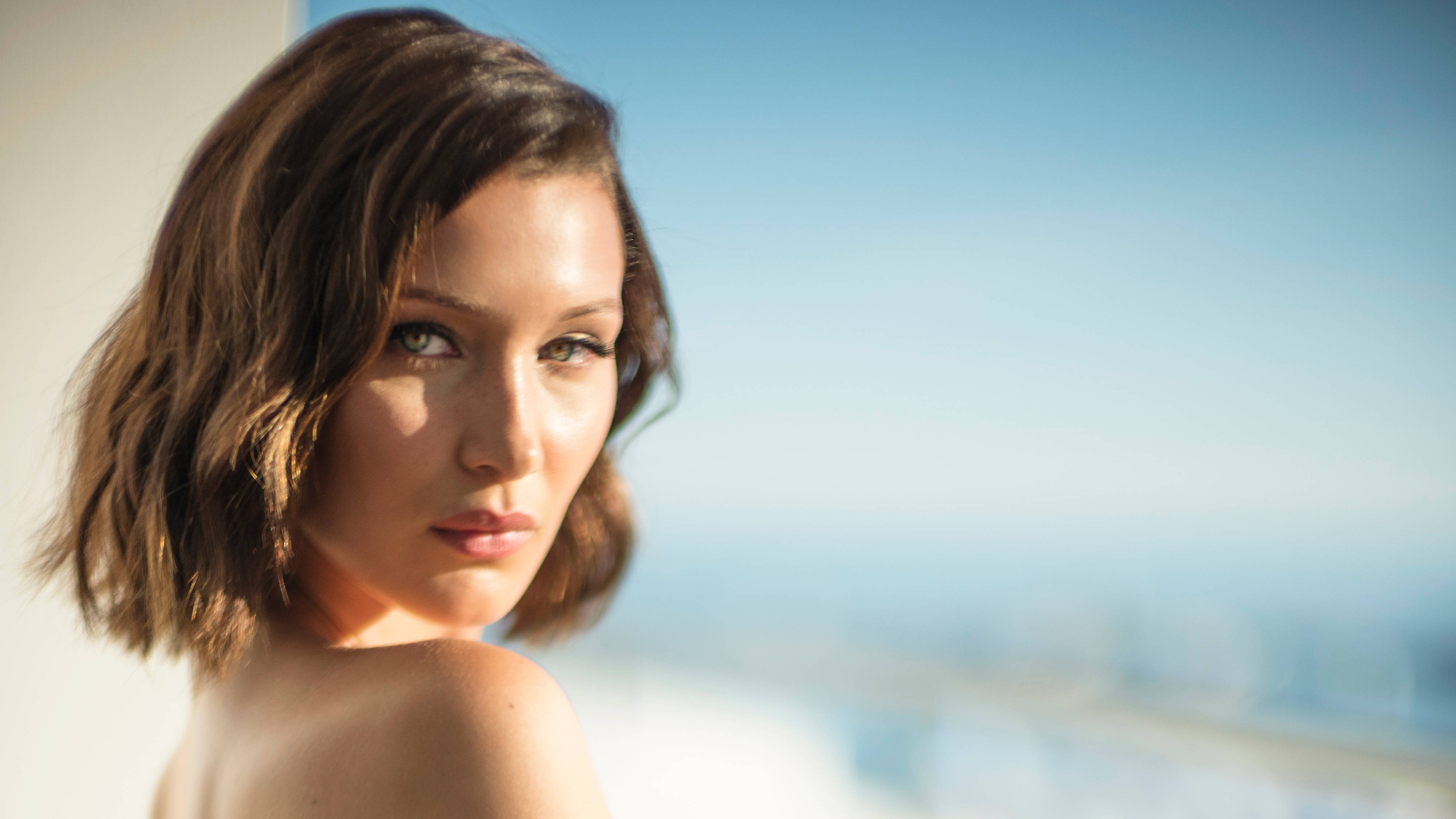 Bella Hadid 桌布 壁紙 — 一位短棕髮女性在明亮的藍天和模糊的城市景觀背景下直視相機。