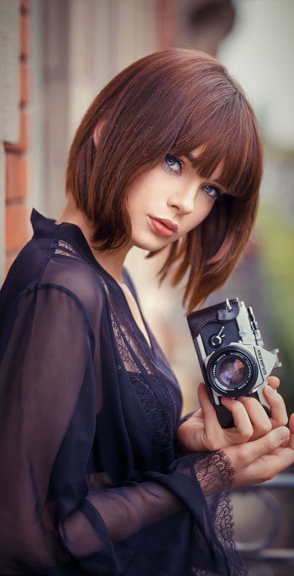 Holding Camera 桌布 壁紙 — 一位短棕髮女性手持復古相機，在柔和自然光中直視觀看者。
