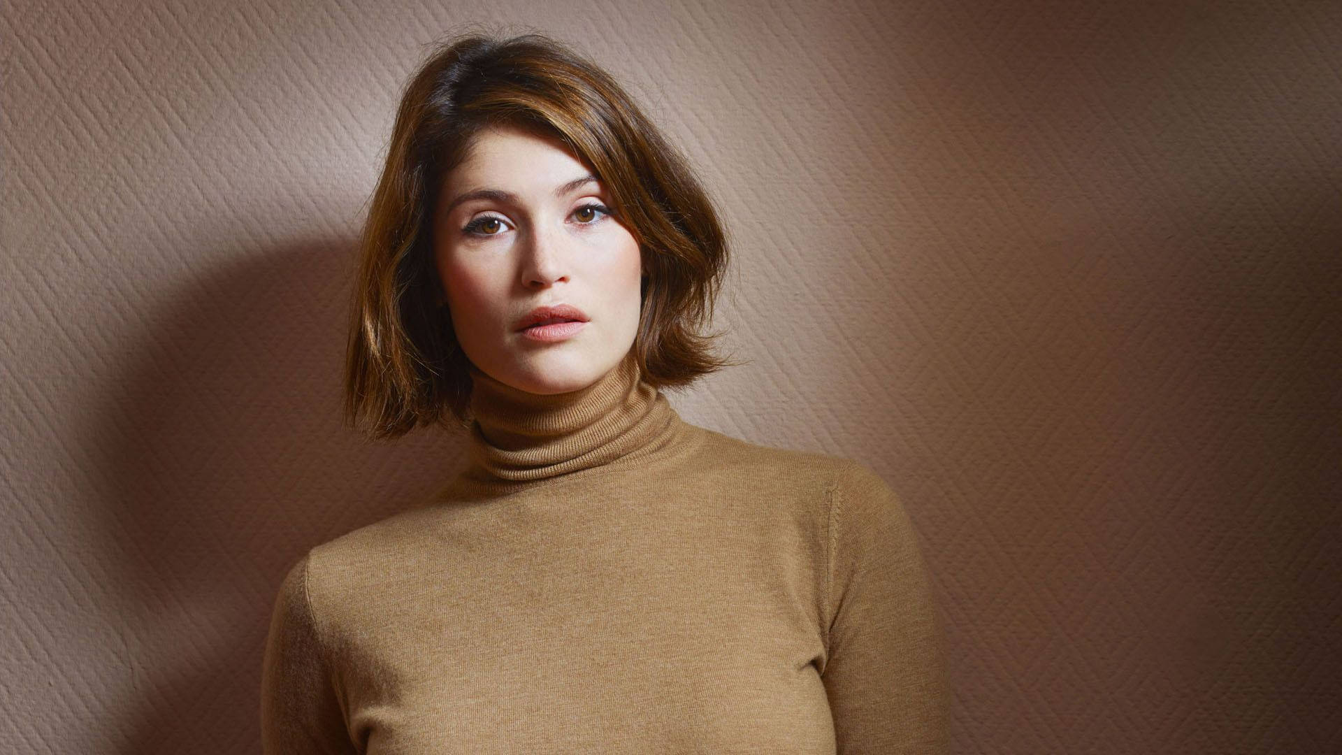 Gemma Arterton 桌布 壁紙 — 一位短棕髮女性穿著棕褐色高領毛衣，背景是帶紋理的米色牆壁，光線柔和。
