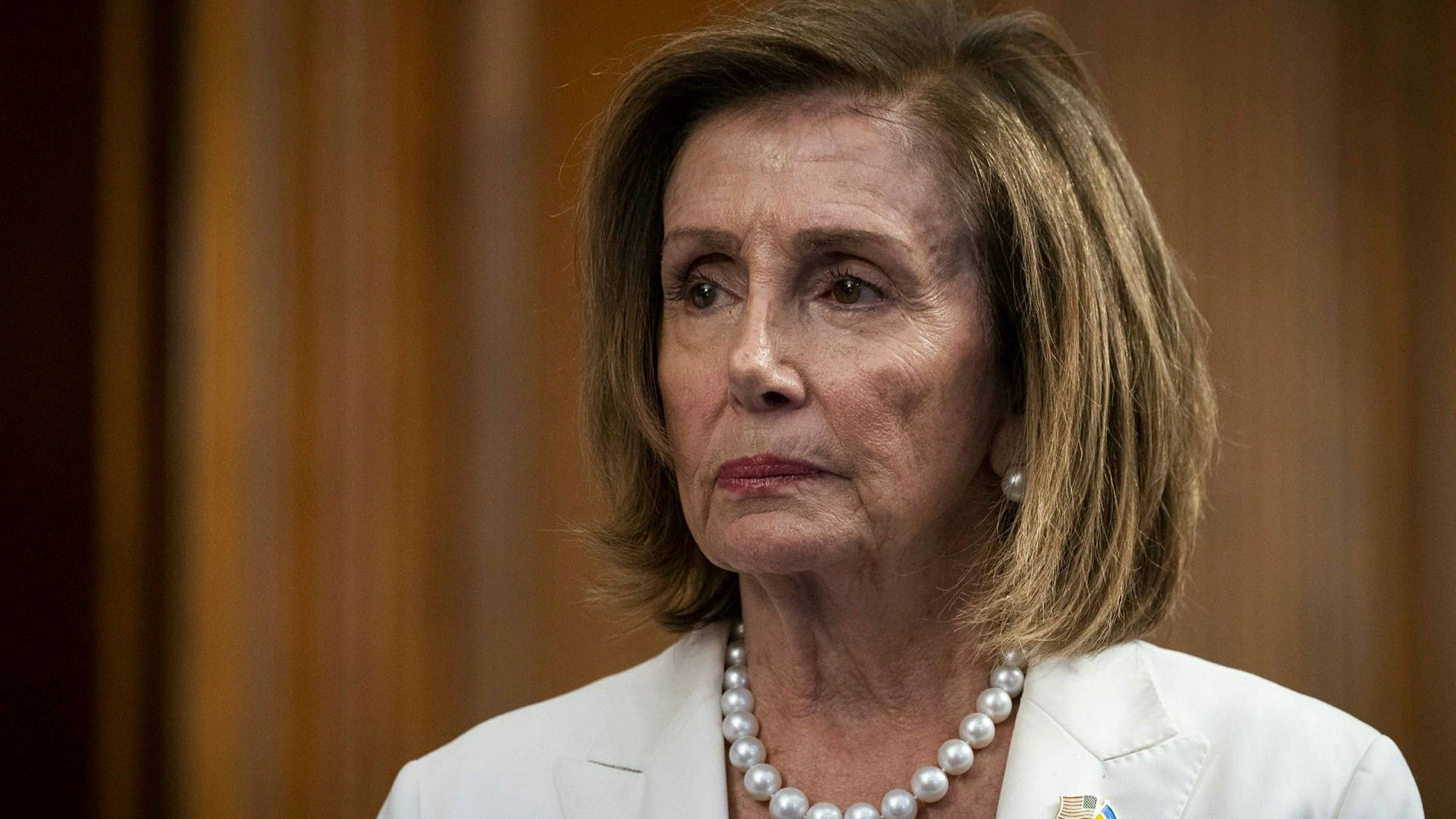 Nancy Pelosi 桌布 壁紙 — 一位短棕髮女性穿著白色西裝外套和珍珠項鍊，以木質背景為襯的特寫肖像。