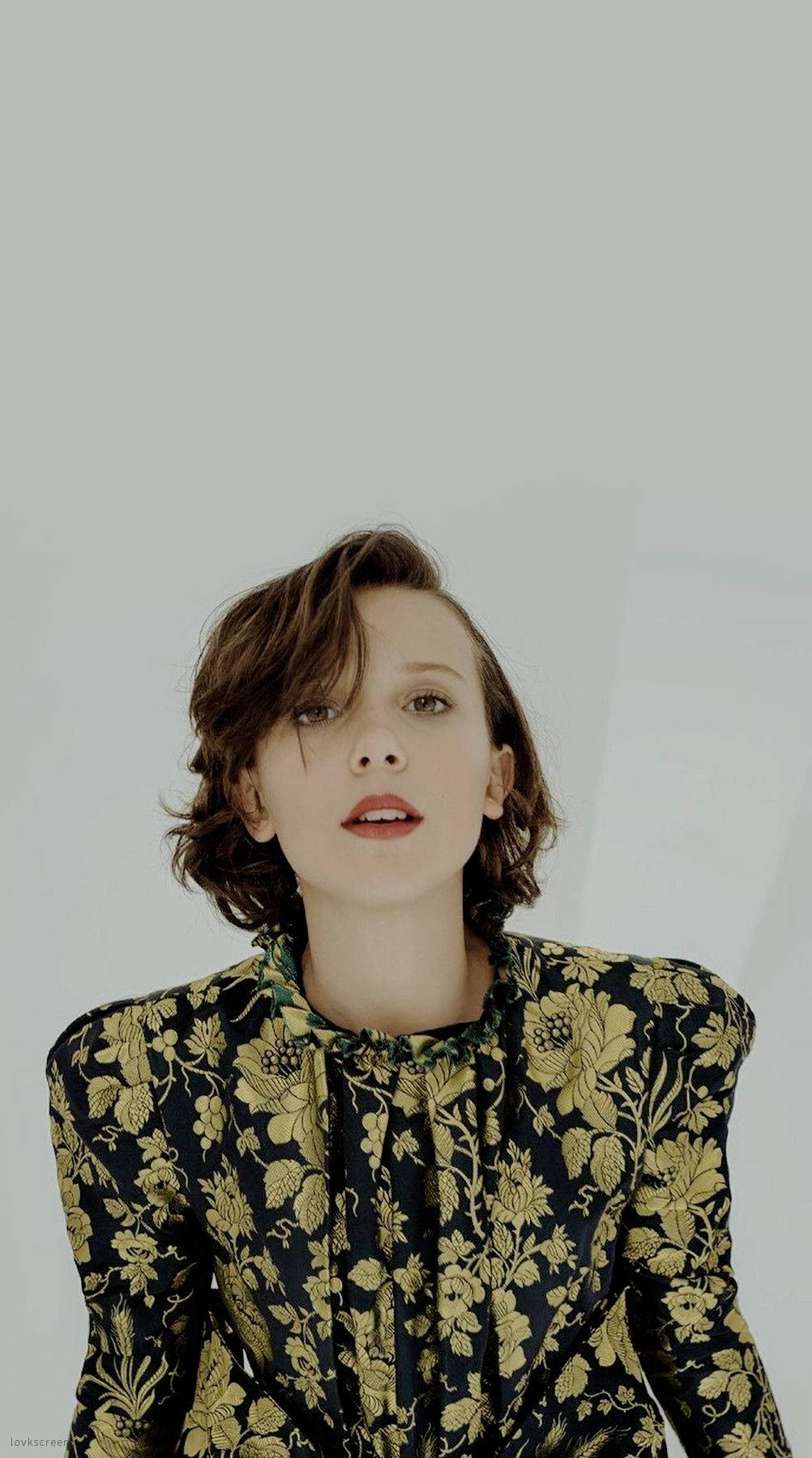 Millie 桌布 壁紙 — 一位短棕髮女性穿著黑色和金色花卉圖案襯衫，背景為純淺灰色。