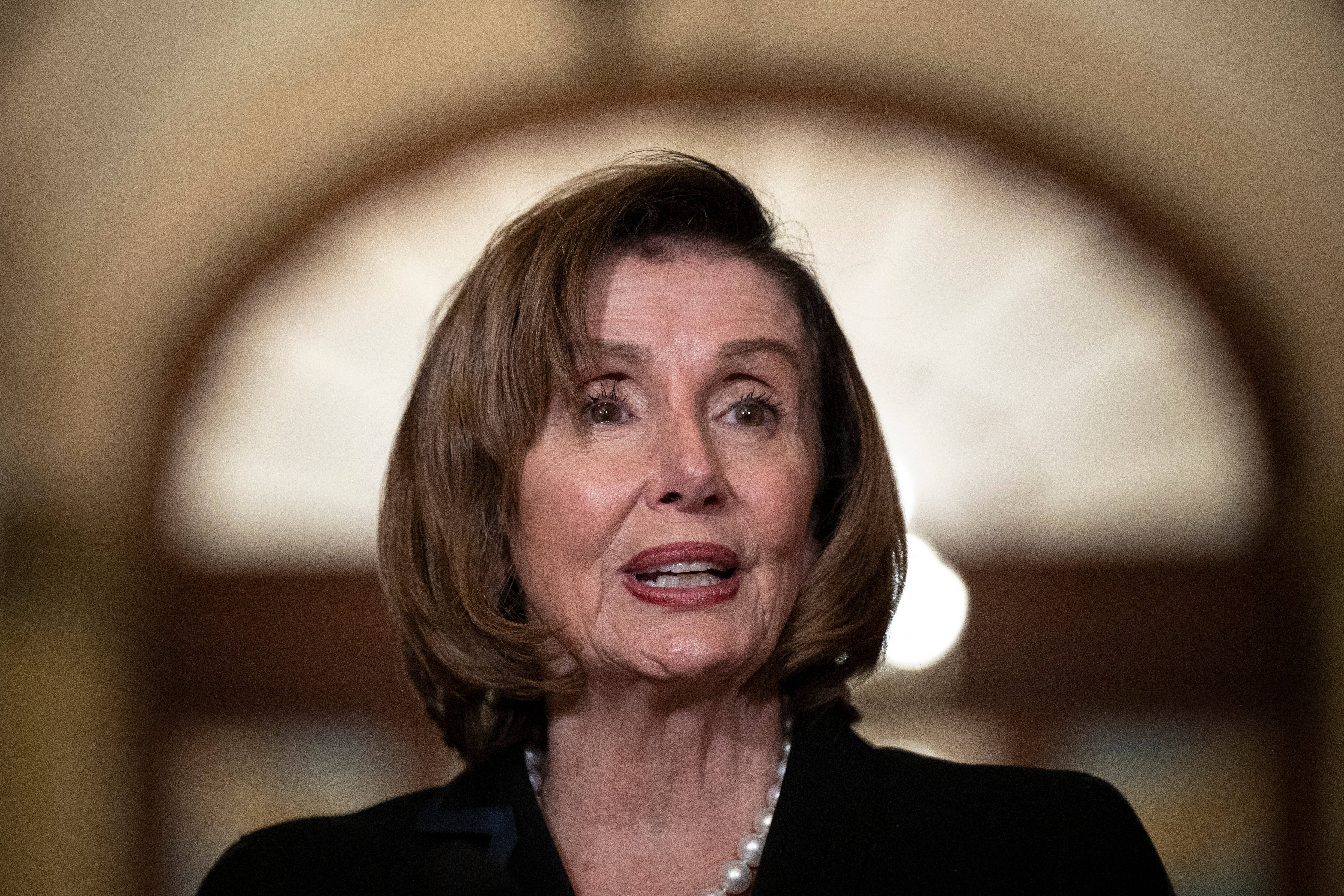 Nancy Pelosi 桌布 壁紙 — 一位短棕髮女性穿著黑色西裝外套和珍珠項鍊的特寫肖像，背景是柔和模糊的拱形。