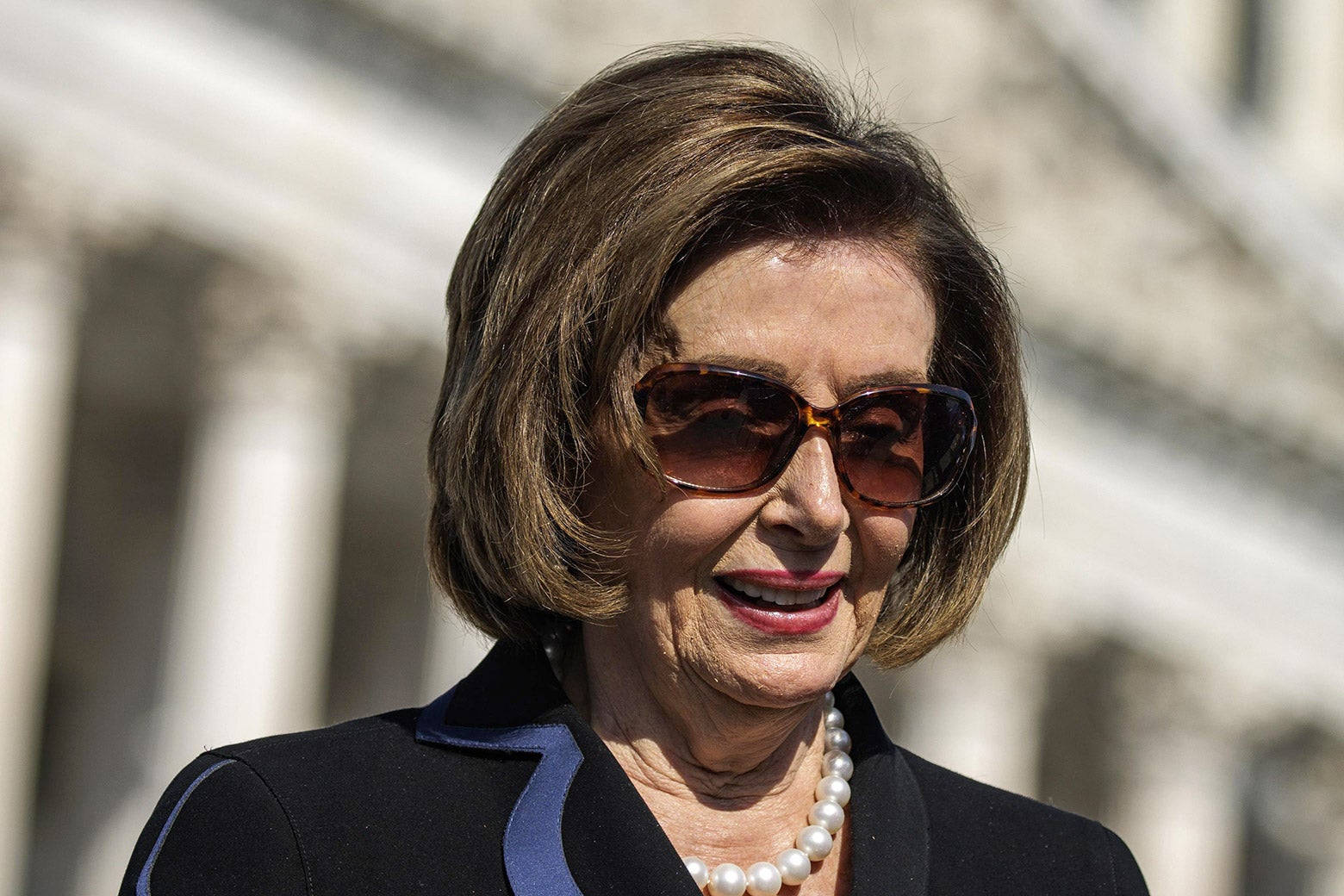 Nancy Pelosi 桌布 壁紙 — 一位短棕髮微笑的女性，戴著太陽眼鏡和珍珠項鍊，背景是模糊的古典建築外牆。