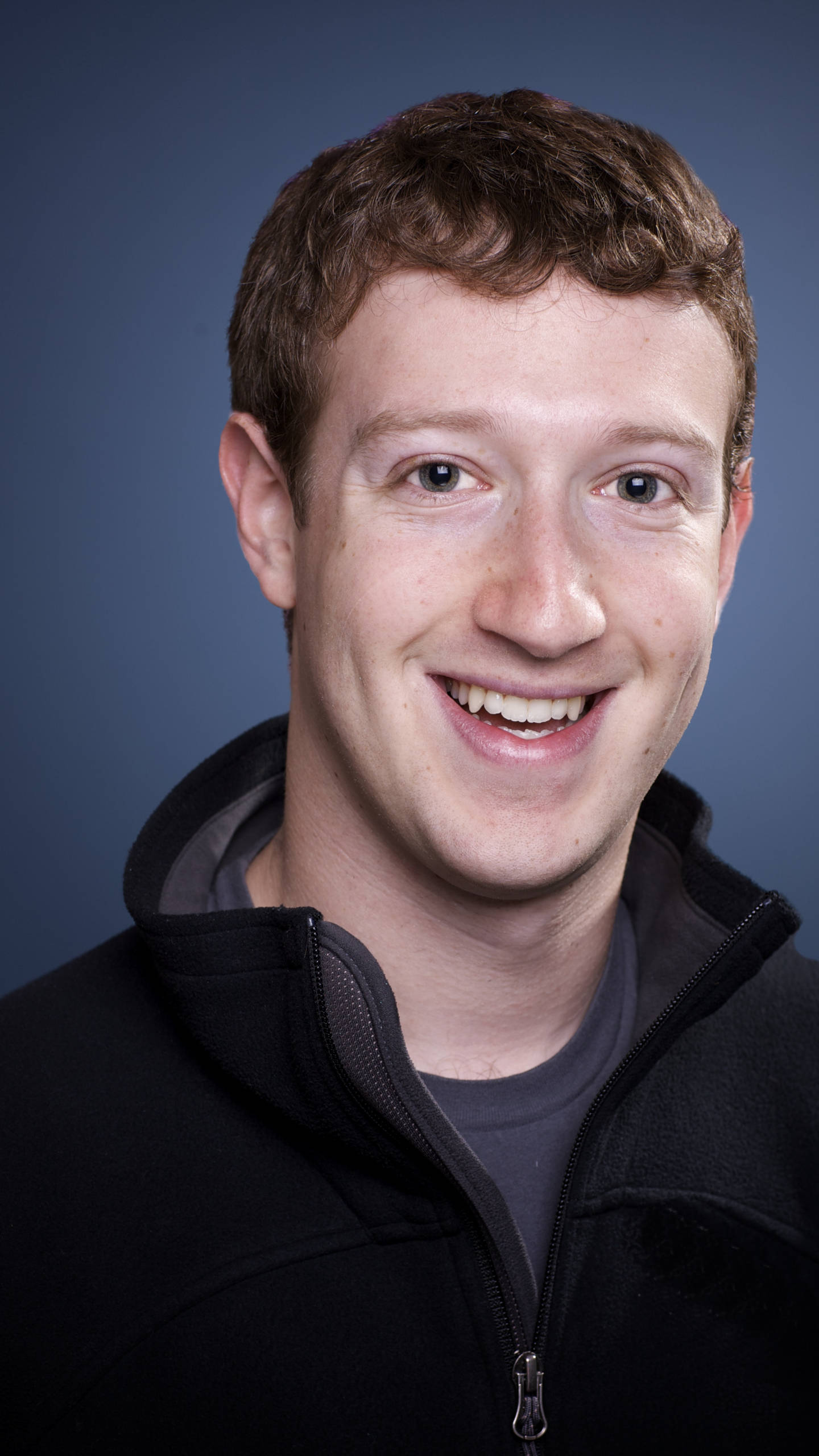 Zuckerberg 桌布 壁紙 — 一位短棕髮微笑的男子，在純藍色背景前穿著黑色拉鍊連帽衫。