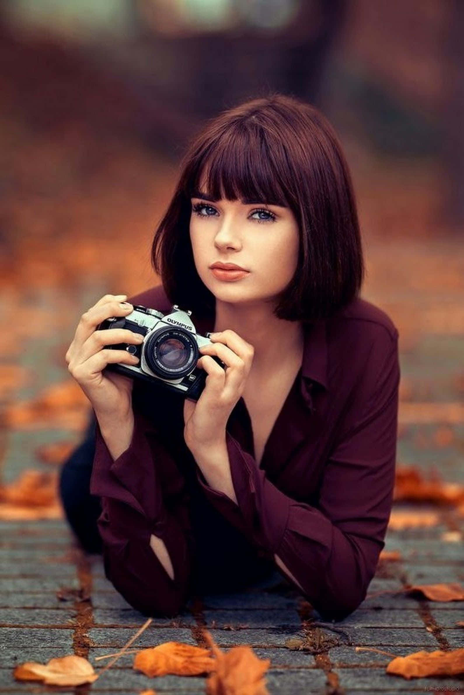 Holding Camera 桌布 壁紙 — 一位短棕髮的年輕女子躺在木棧道上，手持一部復古相機，周圍散落著秋天的落葉。