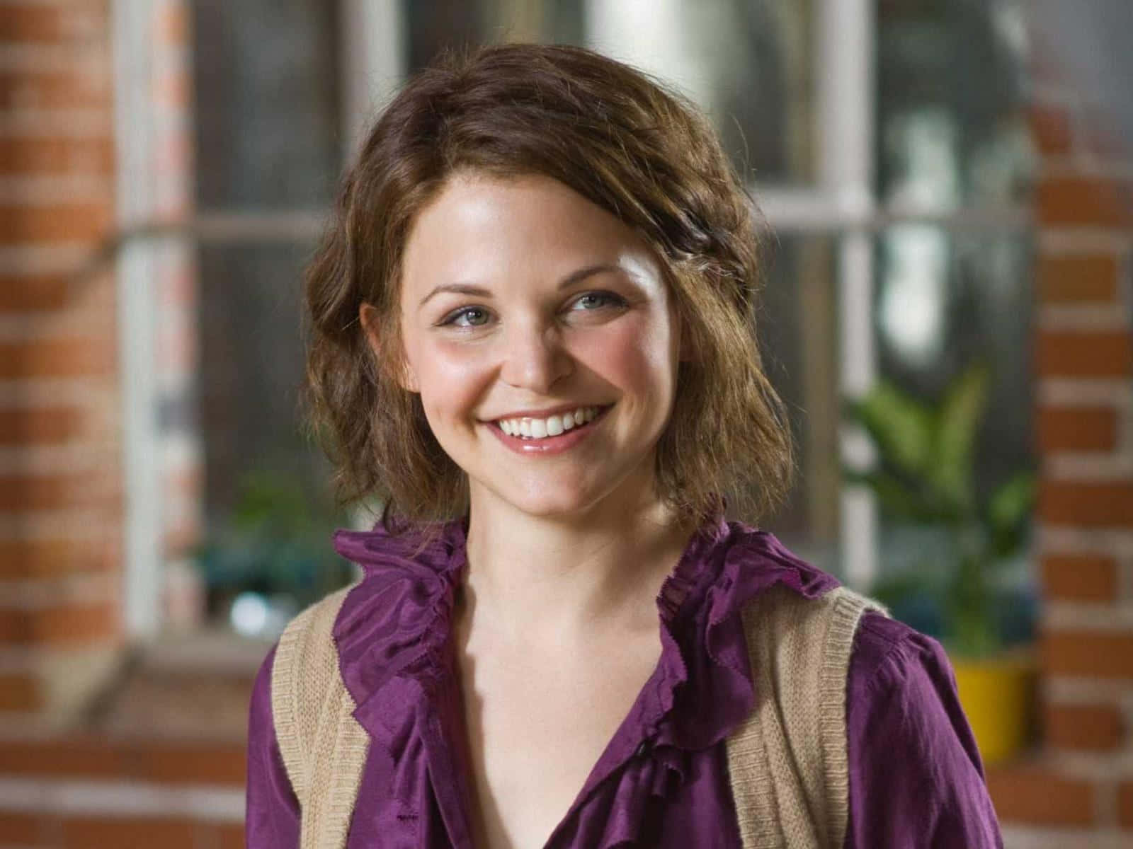ginnifer goodwin 桌布 壁紙 — 一位短棕髮的開朗女性在磚牆和窗戶前溫暖地微笑。