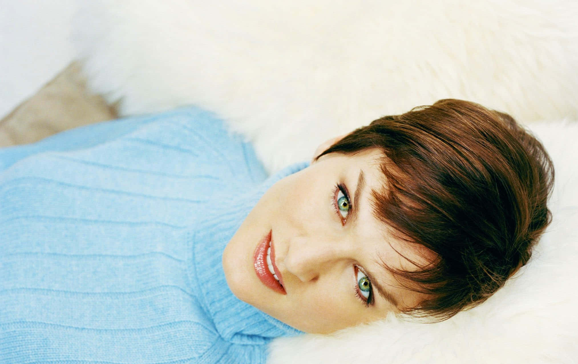 Fiona Bruce 桌布 壁紙 — 一位短棕髮綠眼睛的女性，穿著淺藍色毛衣，躺在蓬鬆的白色表面上微笑。