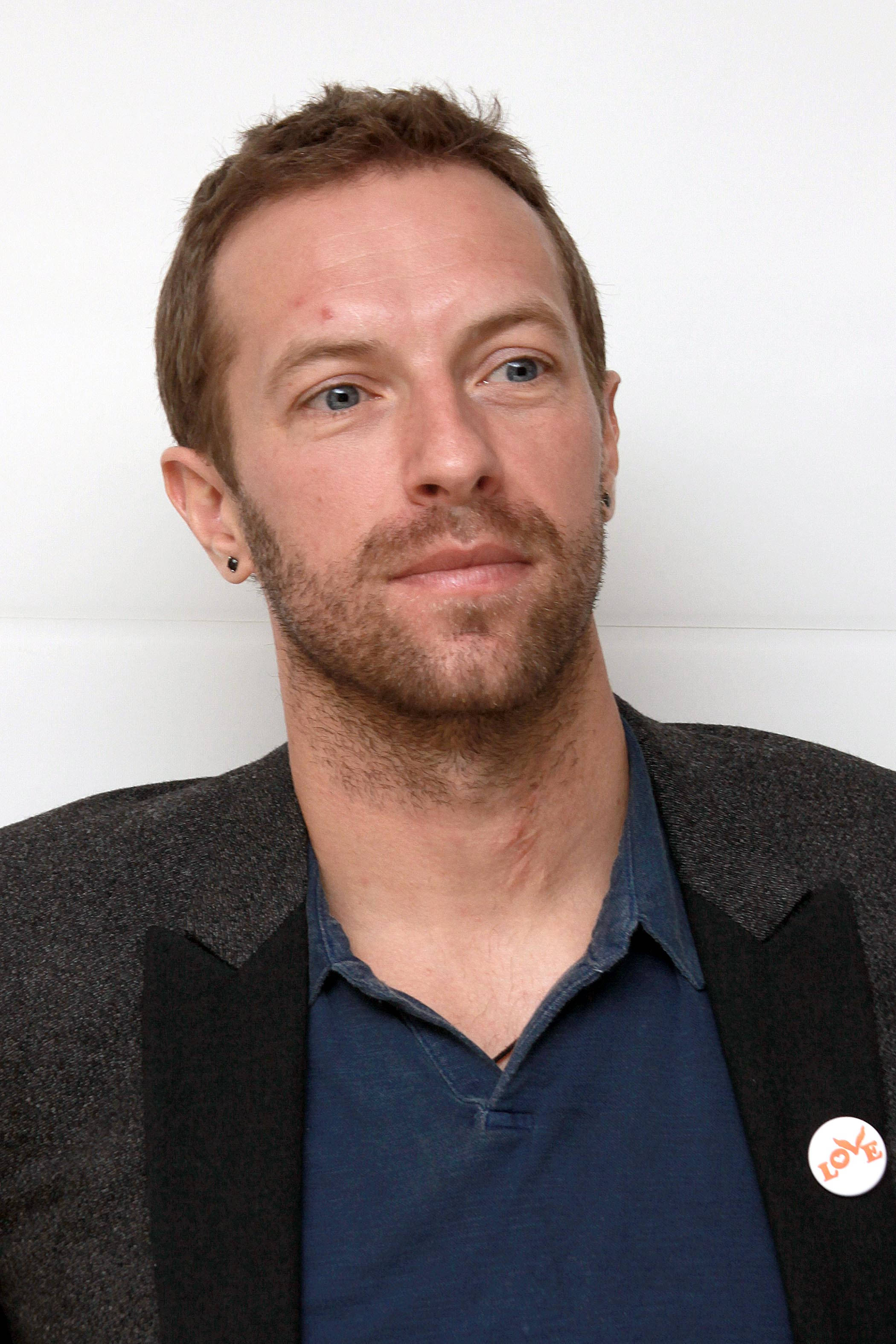 Chris Martin 桌布 壁紙 — 一位短棕髮藍眼的男士，在純白背景前穿著深色西裝外套內搭藍色襯衫。