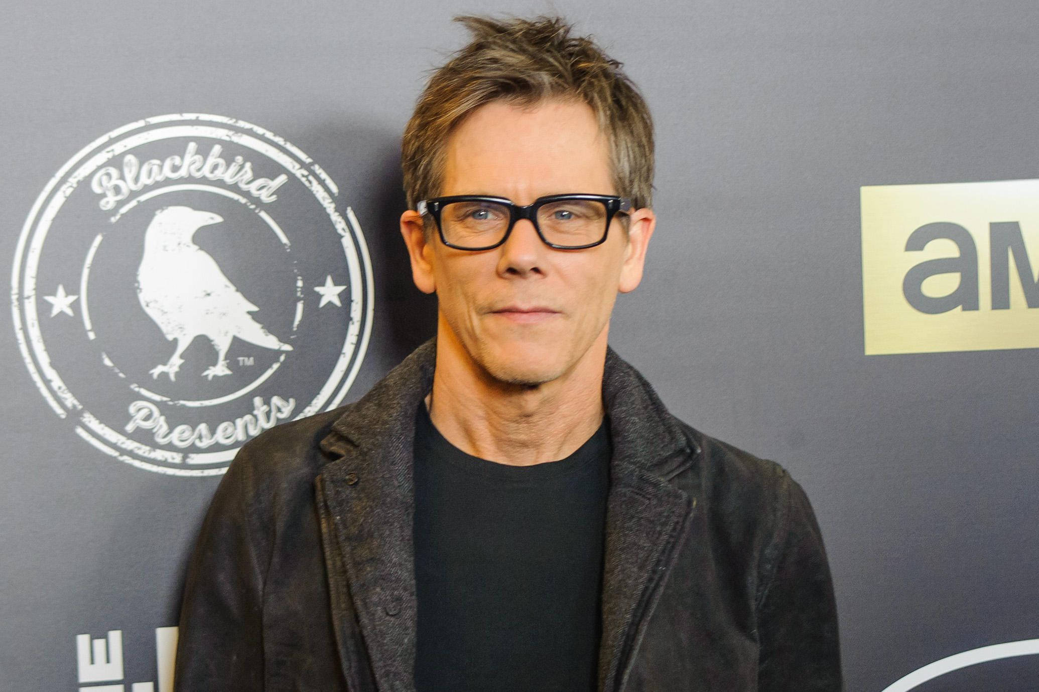 actor 桌布 壁紙 — 一位短灰髮、戴黑框眼鏡的男子，在灰色背景前穿著黑色襯衫外搭深色外套，背景上有標誌。