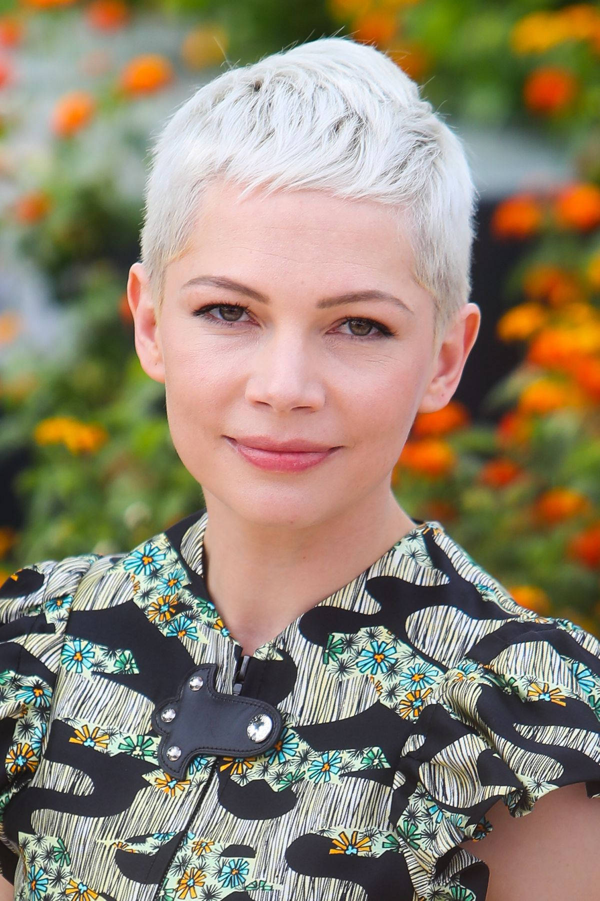 Celebrity 桌布 壁紙 — 一位短白金髮女性在充滿活力的橙綠色花卉背景下溫柔微笑。