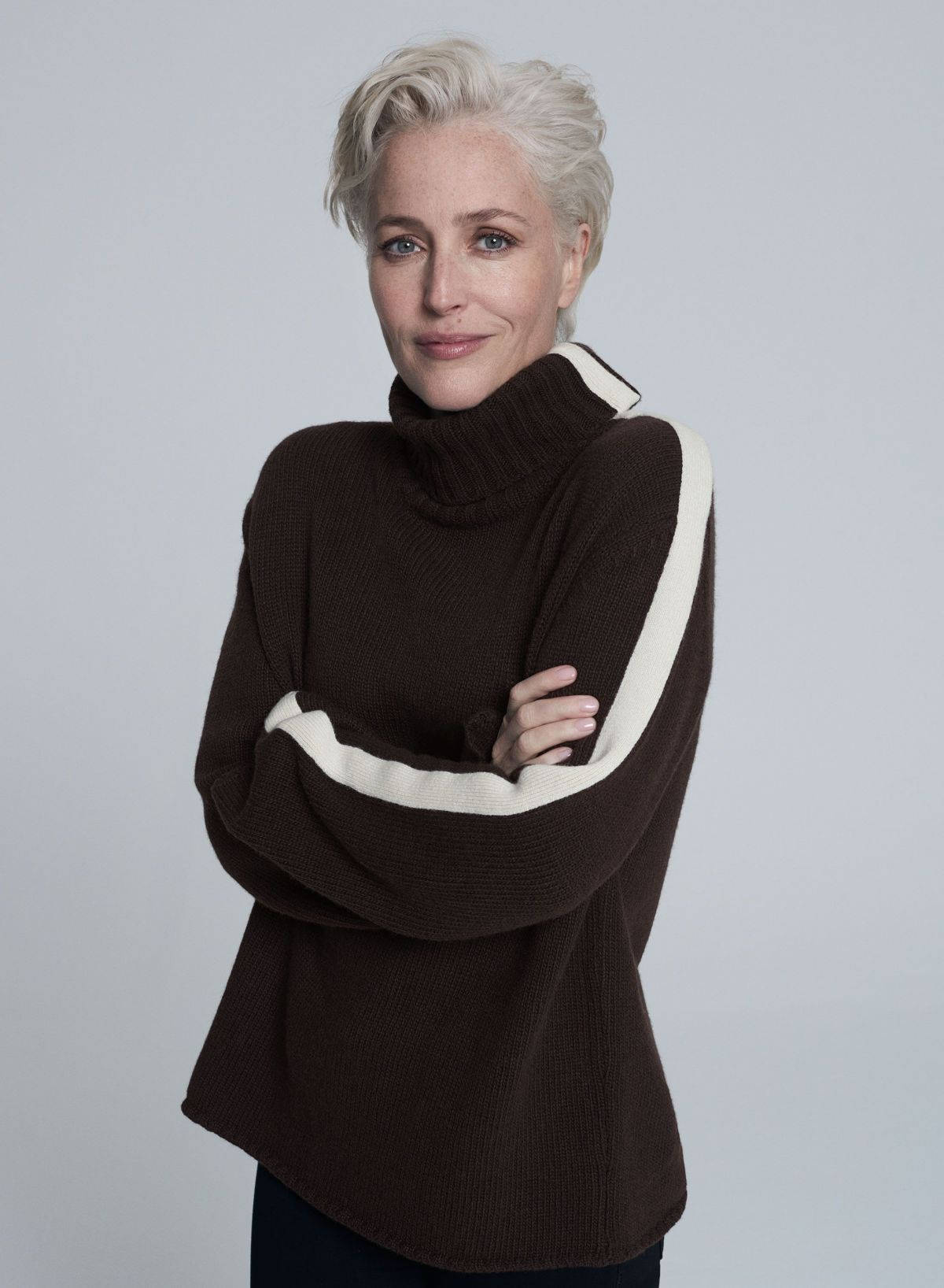 gillian anderson 桌布 壁紙 — 一位短白金髮女性穿著帶有奶油色條紋的棕色高領毛衣，站在純灰色背景前。