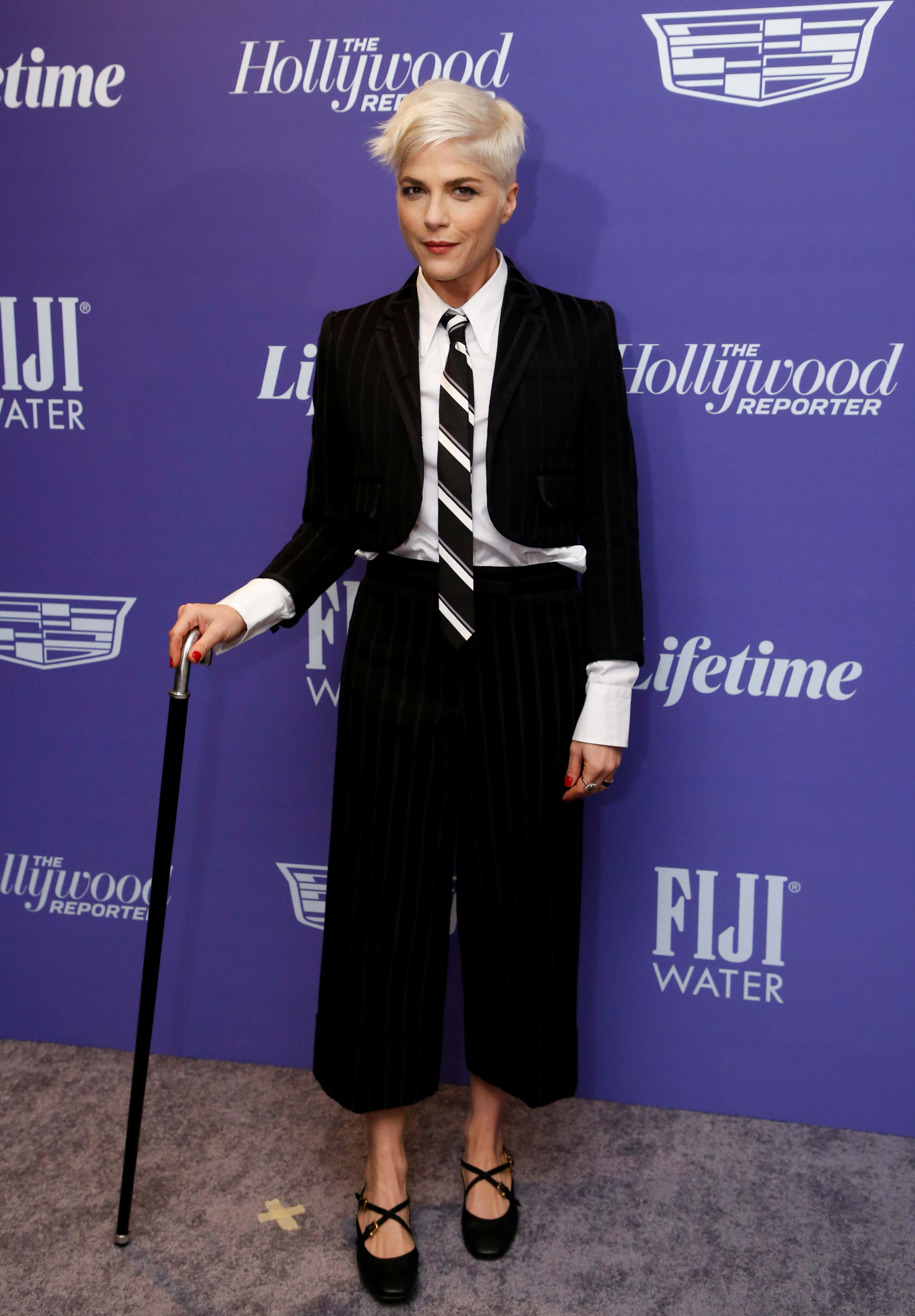 Selma Blair 桌布 壁紙 — 一位短白金髮的女性自信地穿著黑色細條紋西裝，在紫色品牌背景前拿著手杖。